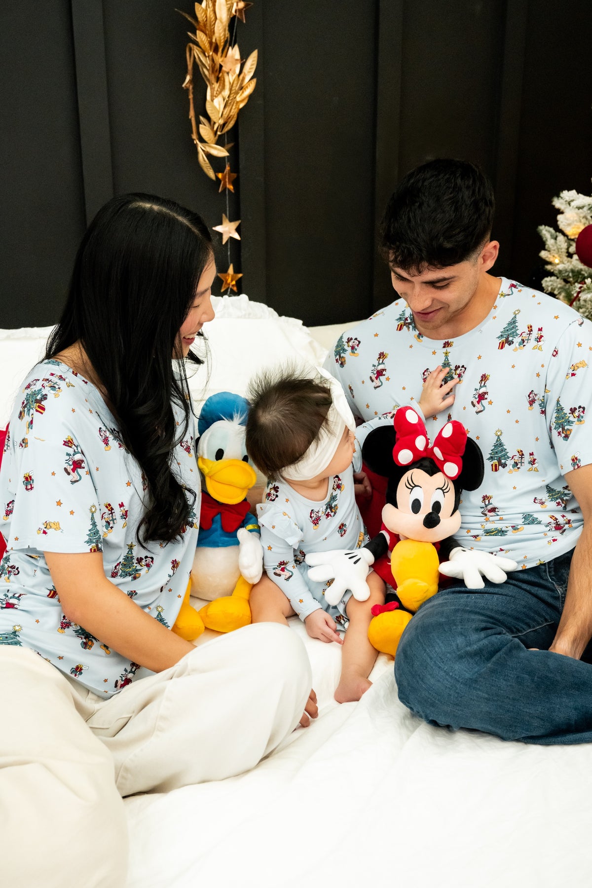 A family of three wearing matching light blue Disney-themed apparel sits together on a white bed, surrounded by Disney plush toys including Donald Duck, Minnie Mouse, and Mickey Mouse.