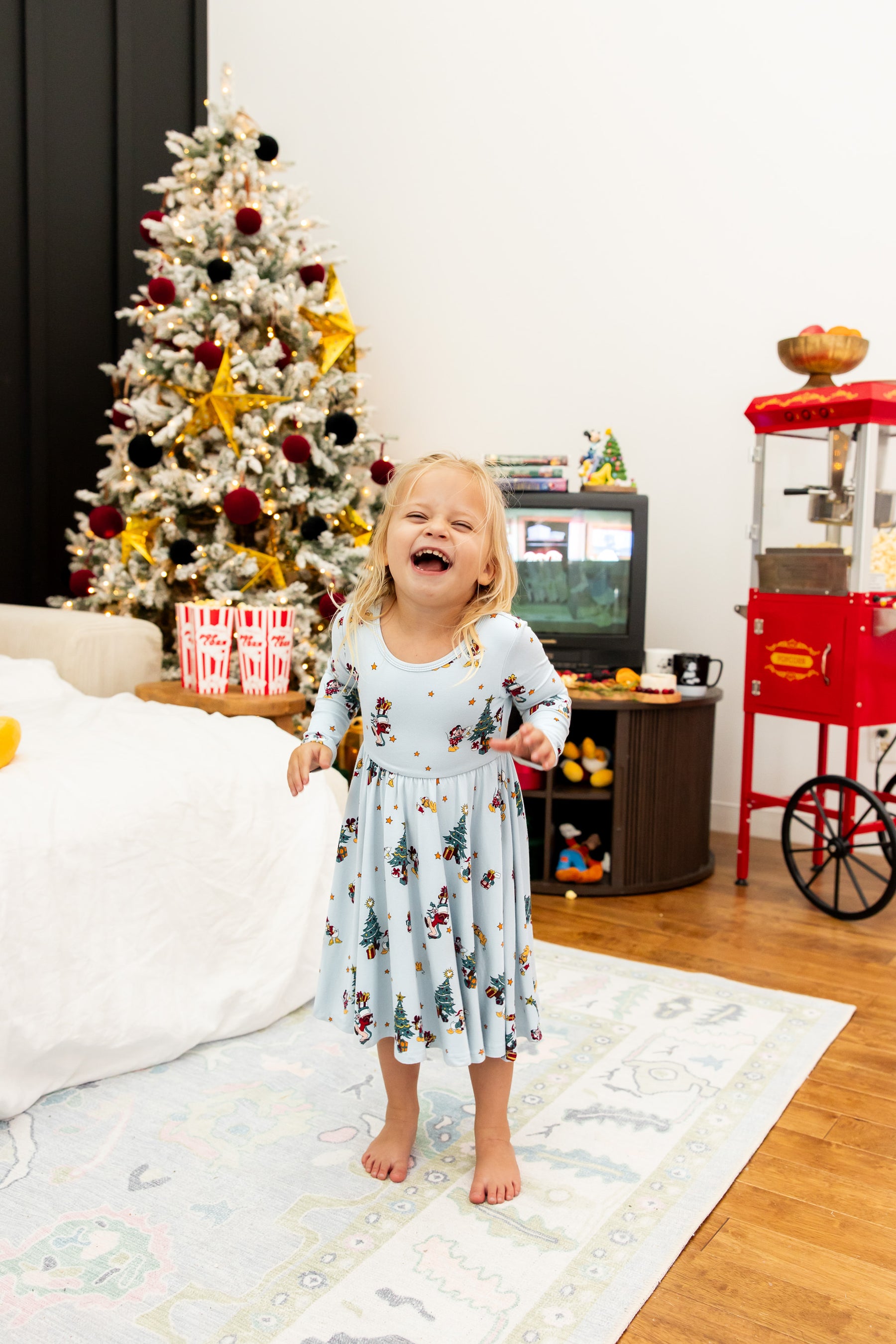 Girl laughing in Long Sleeve Twirl Dress in Mickey & Friends Christmas print standing in a living room with Christmas tree in the background