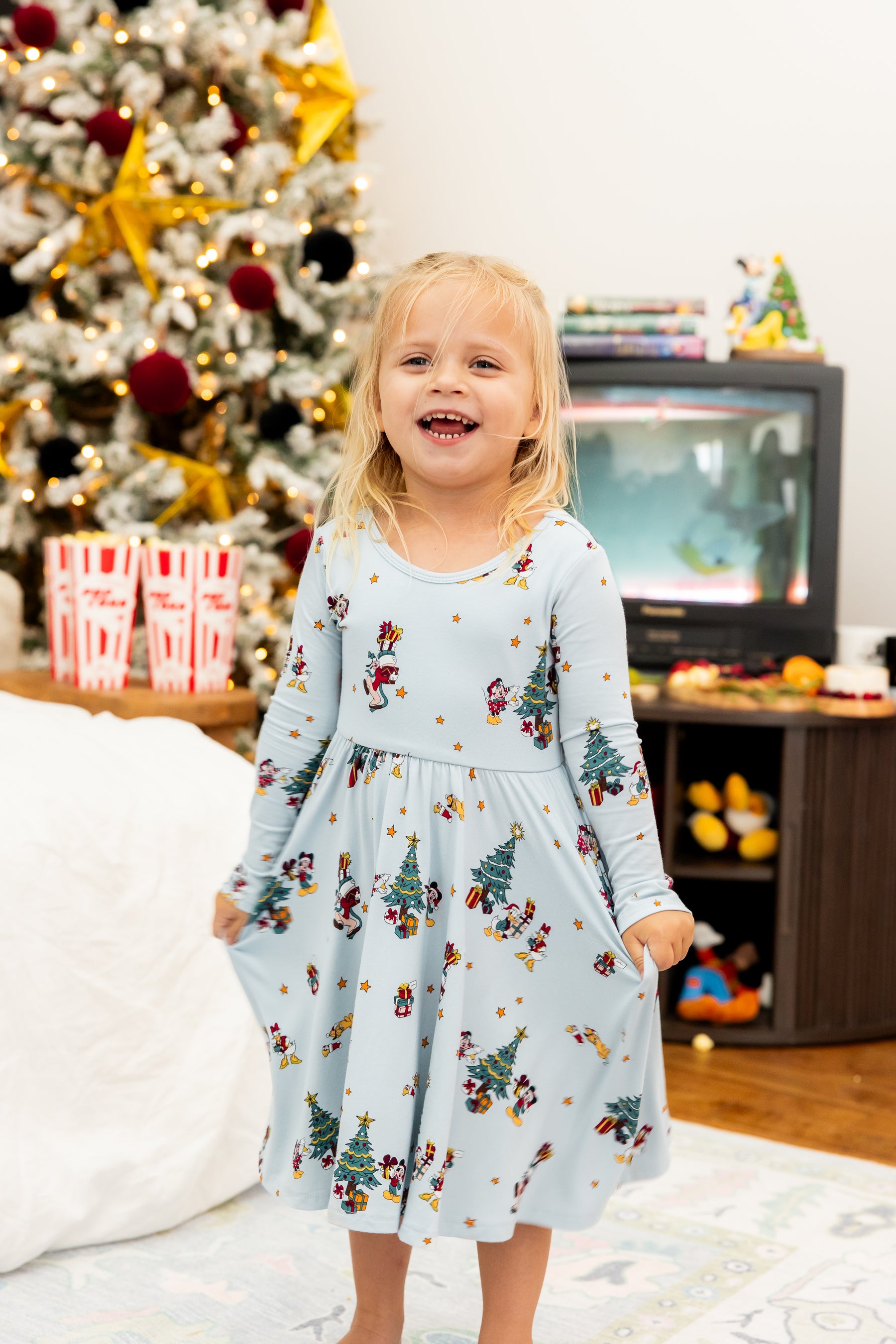 Girl wearing Long Sleeve Twirl Dress in Mickey & Friends Christmas print standing in a living room with Christmas tree in the background