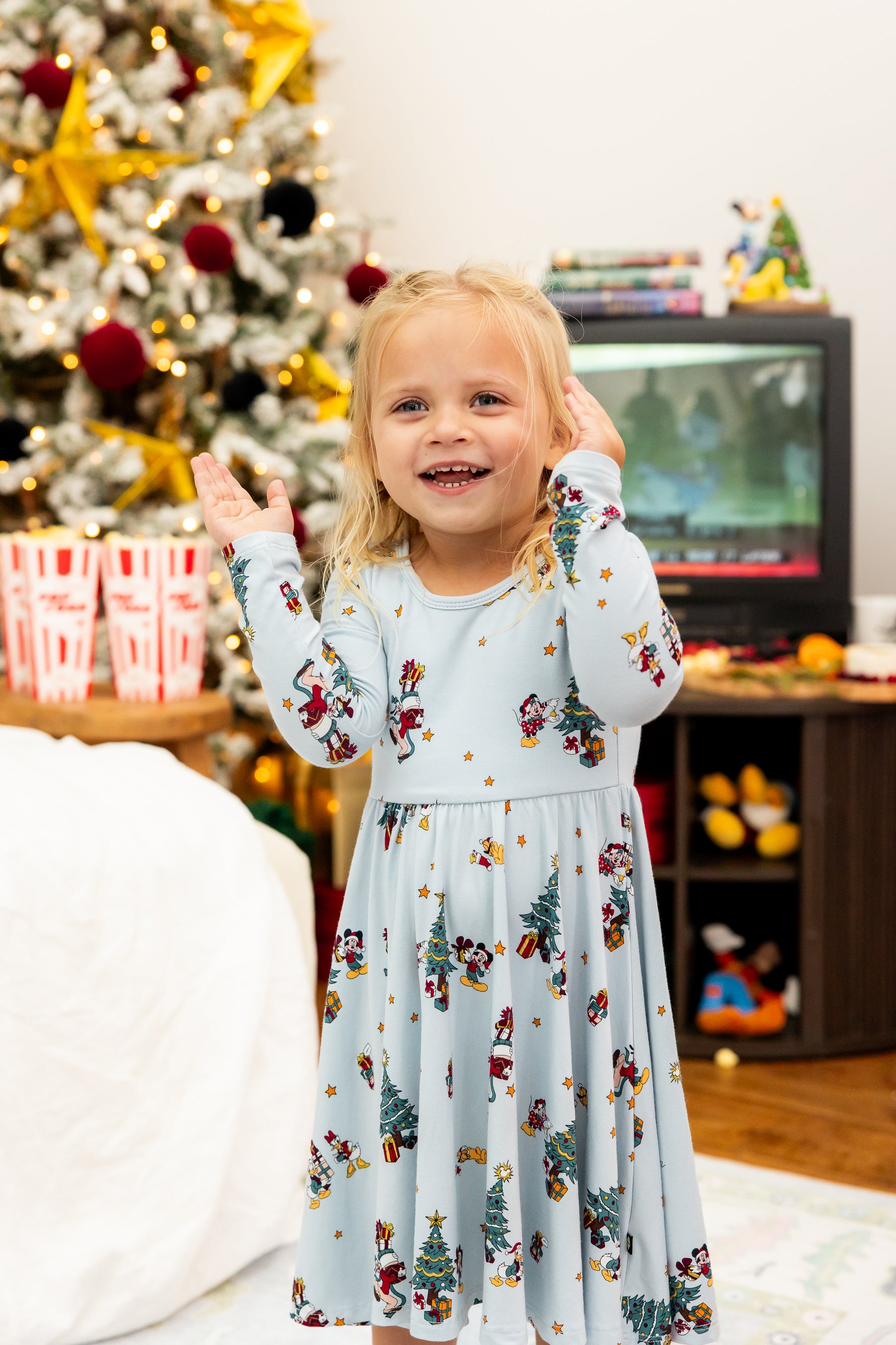 Girl wearing Long Sleeve Twirl Dress in Mickey & Friends Christmas print standing in a living room with Christmas tree in the background