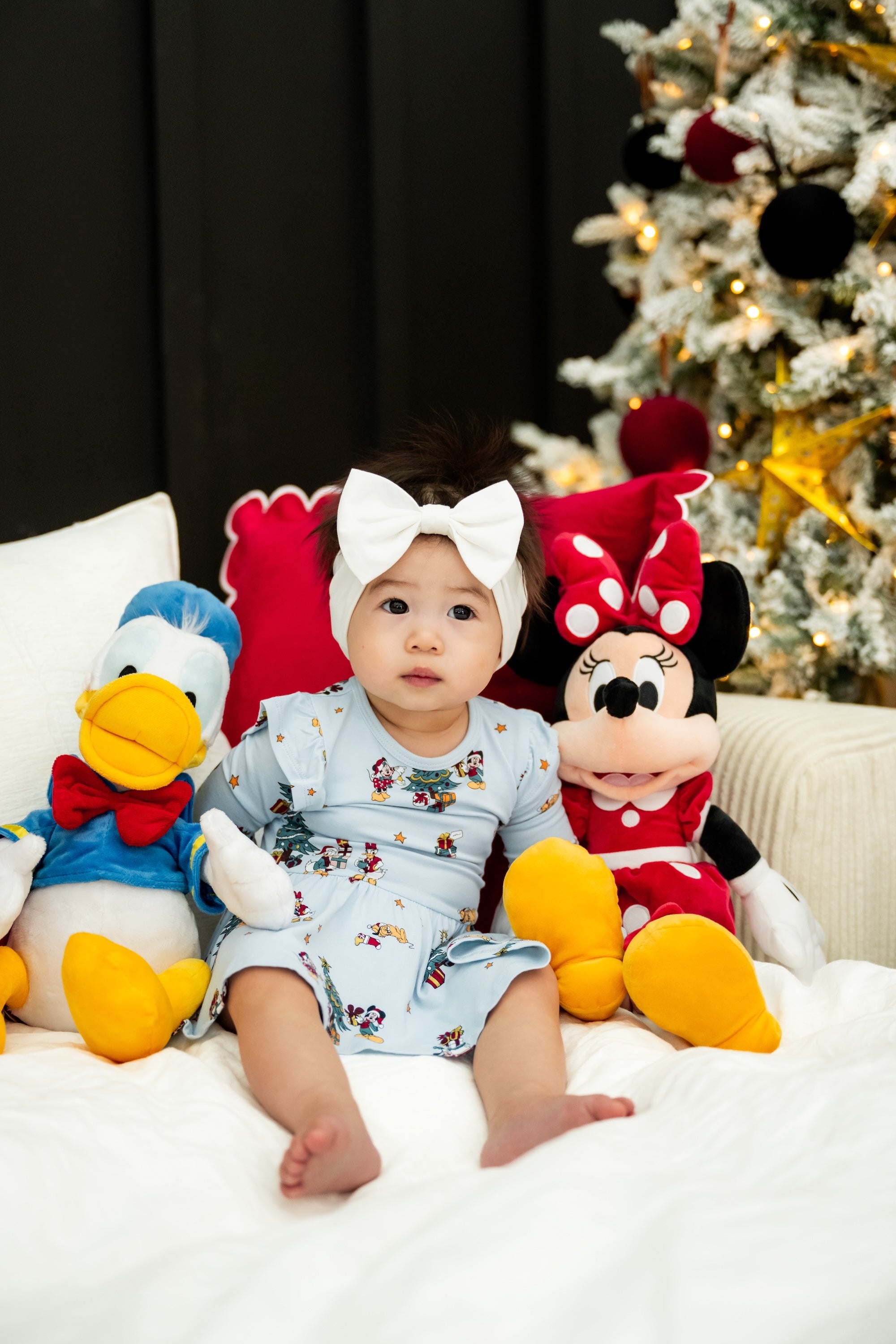 Baby wearing Long Sleeve Twirl Bodysuit Dress in Mickey & Friends Christmas print sitting with stuff toy on a white sofa with Christmas tree in the back