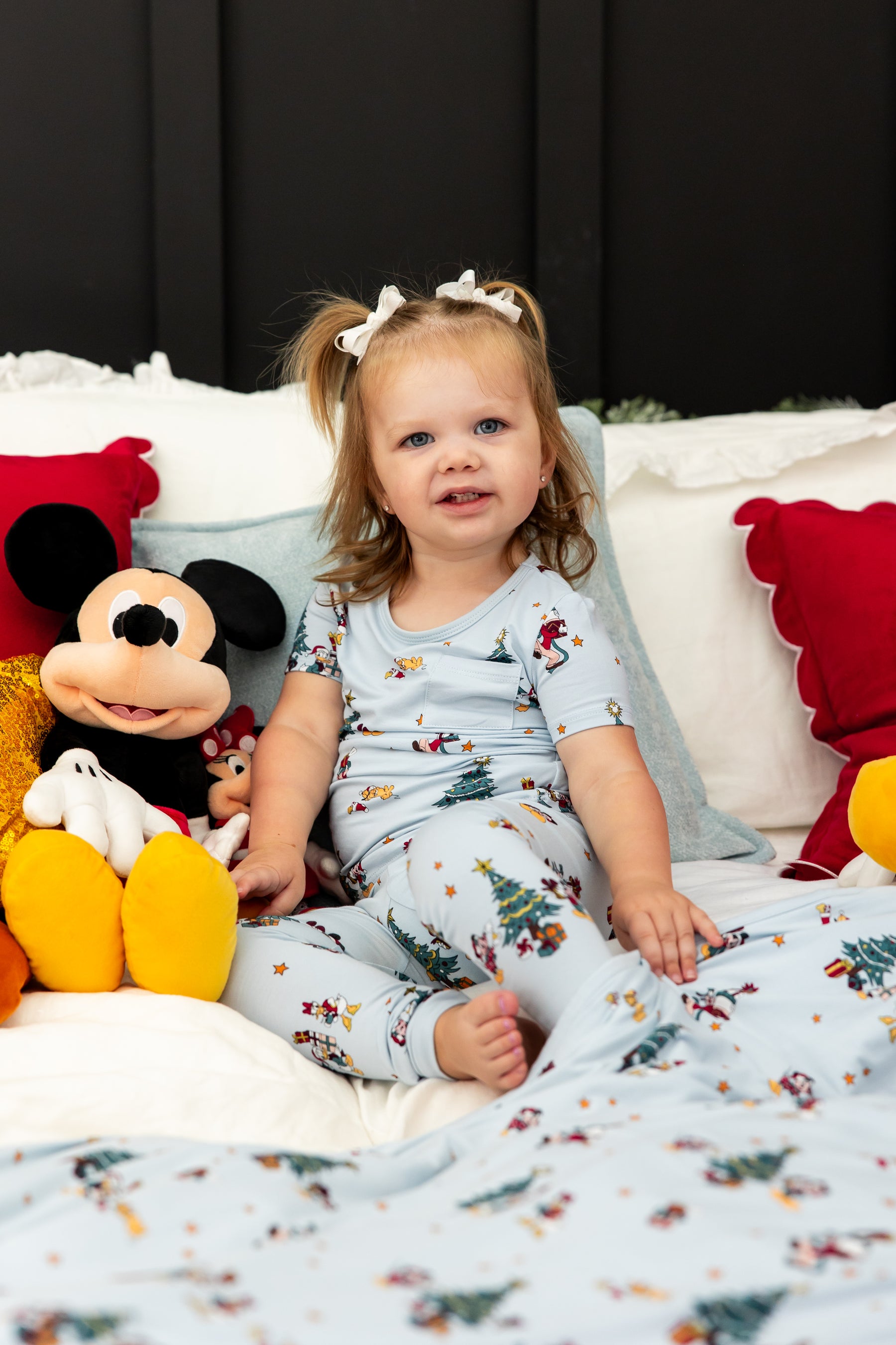 Girl wearing Short Sleeve with Pants Pajamas in Mickey & Friends Christmas print sitting on bed with stuff toys