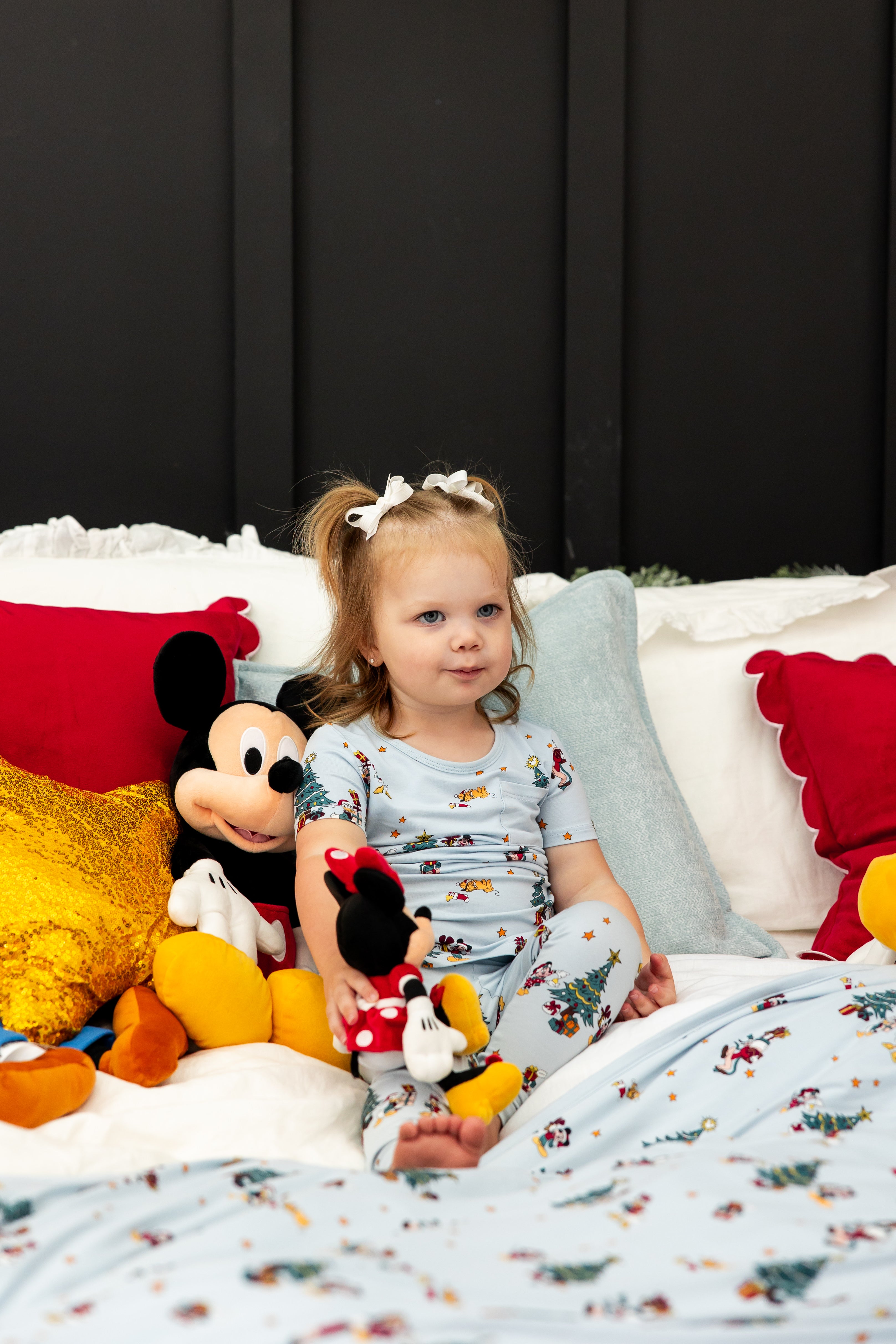 Girl wearing Short Sleeve with Pants Pajamas in Mickey & Friends Christmas print sitting on bed with stuff toys
