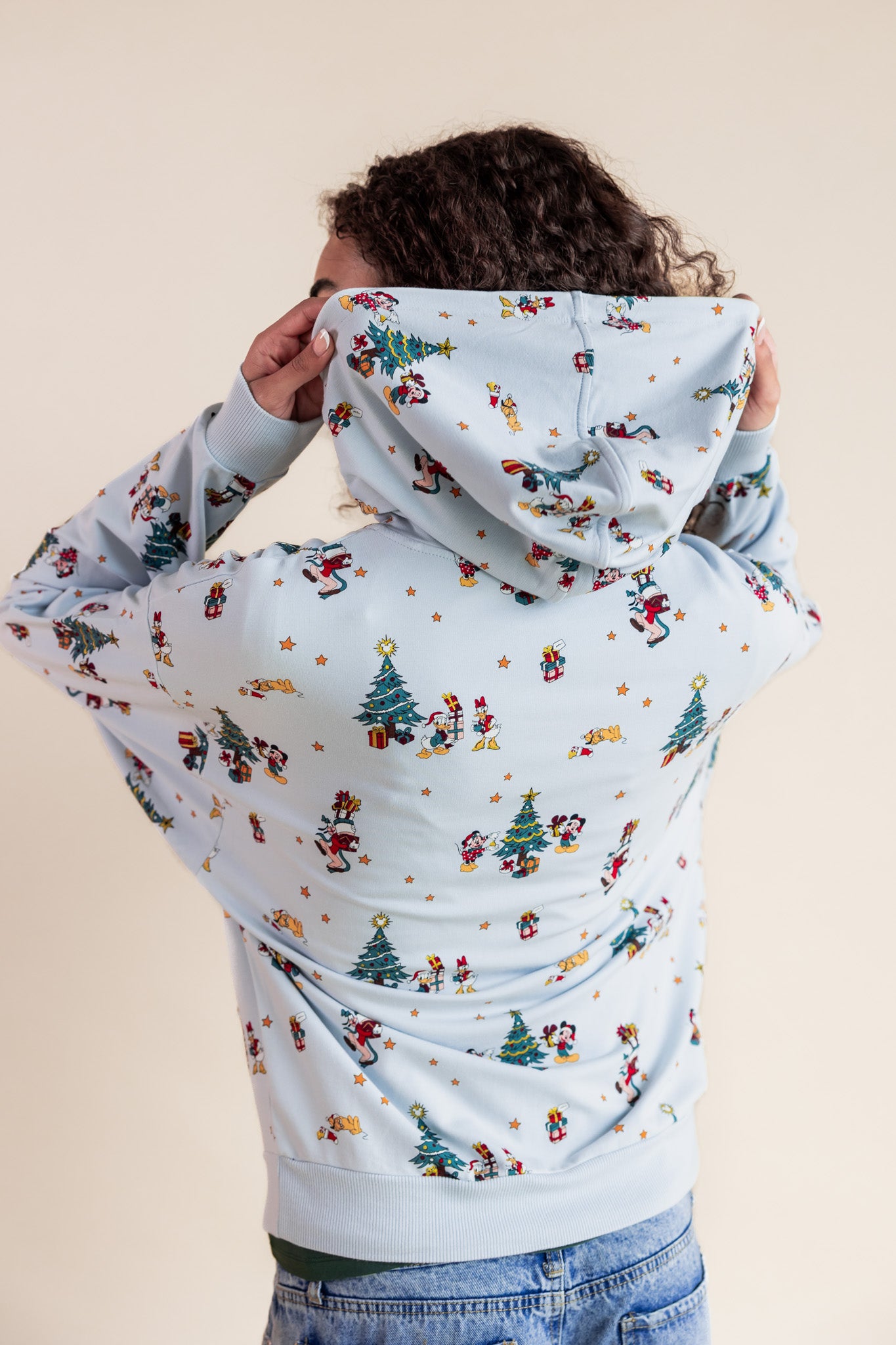 Woman showcasing the hoodie of Women's Hoodie in Mickey & Friends Christmas print over blue jeans on a beige background