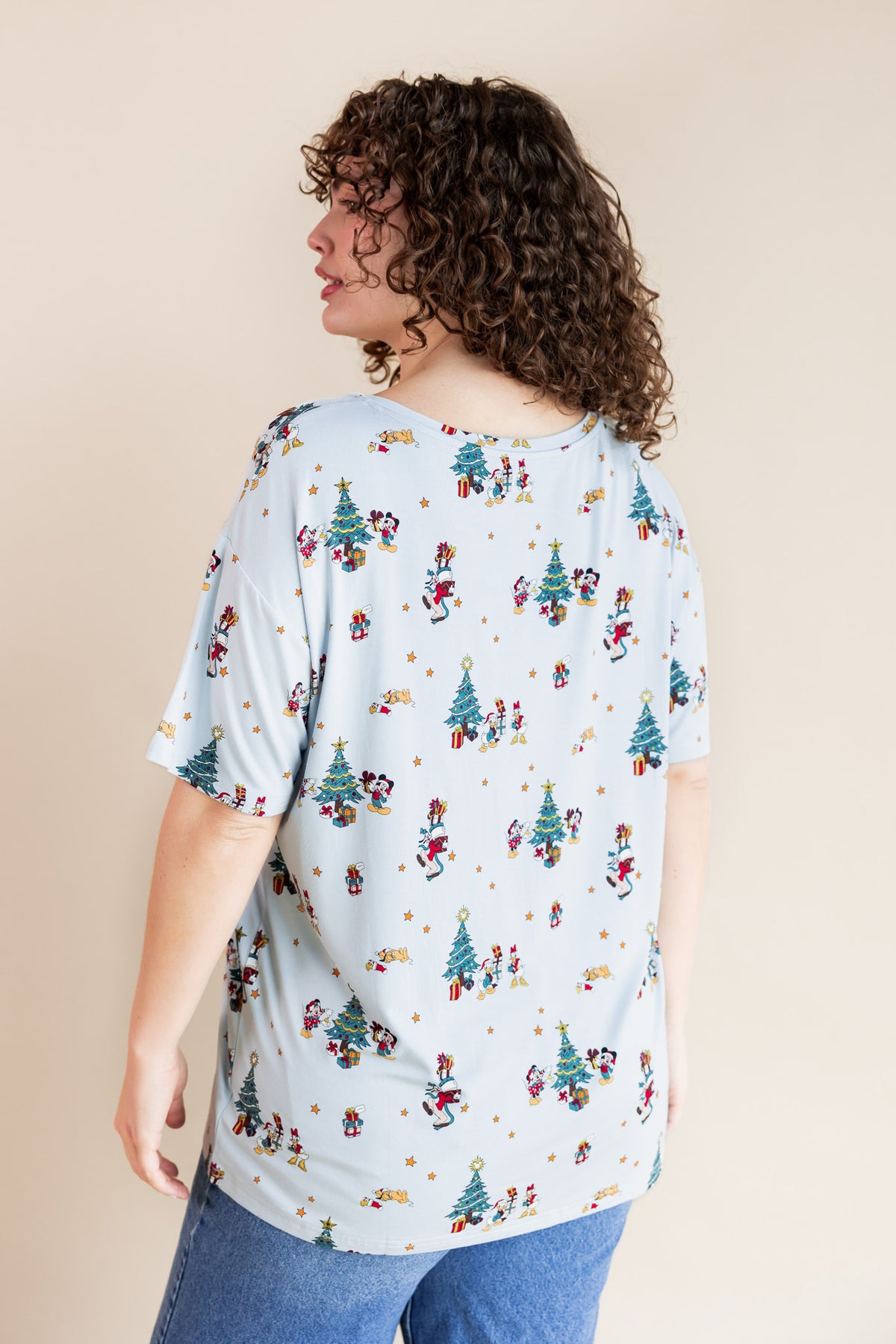 Back side of Woman wearing Women's Oversized V-Neck Tee in Mickey & Friends Christmas print over blue jeans on a beige background