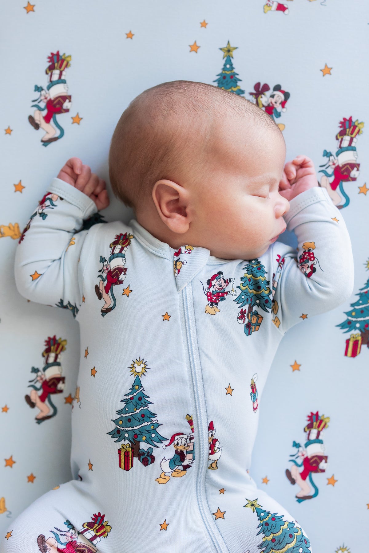 Baby sleeping wearing Zippered Footie in Mickey & Friends Christmas print over similar print sheet