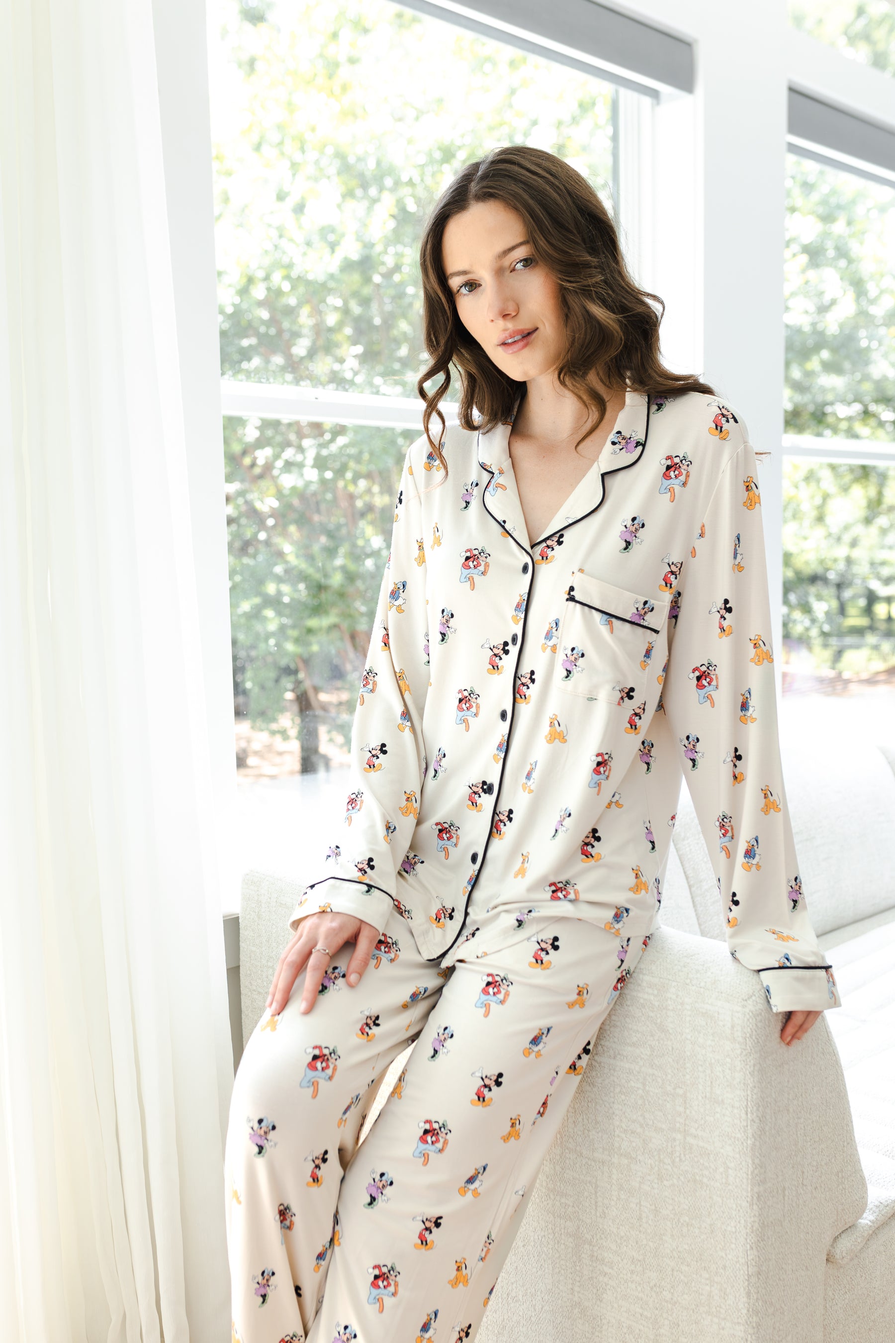 Model posing in Long-Sleeved Women's Pajama Set in Mickey Mouse & Friends print standing against a white sofa in living room 