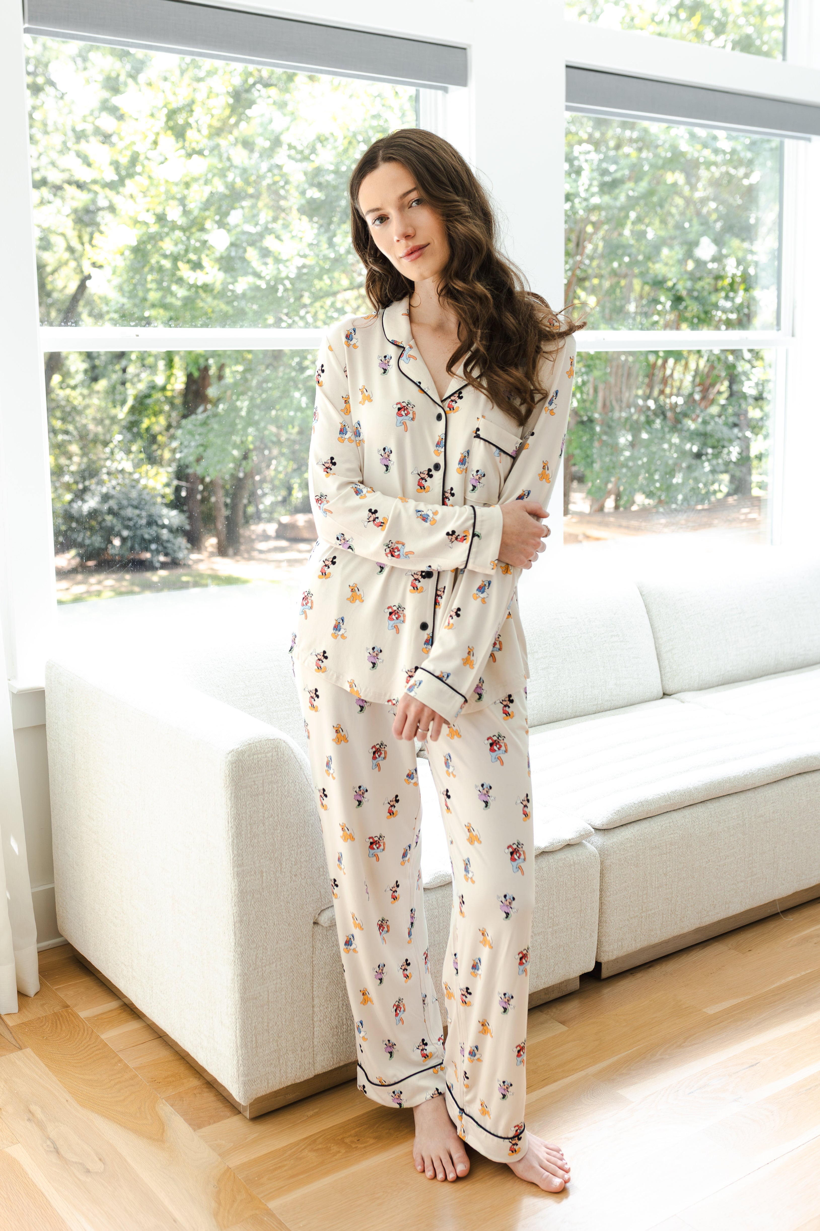 Model wearing Long-Sleeved Women's Pajama Set in Mickey Mouse & Friends print standing in front of a white sofa in living room 