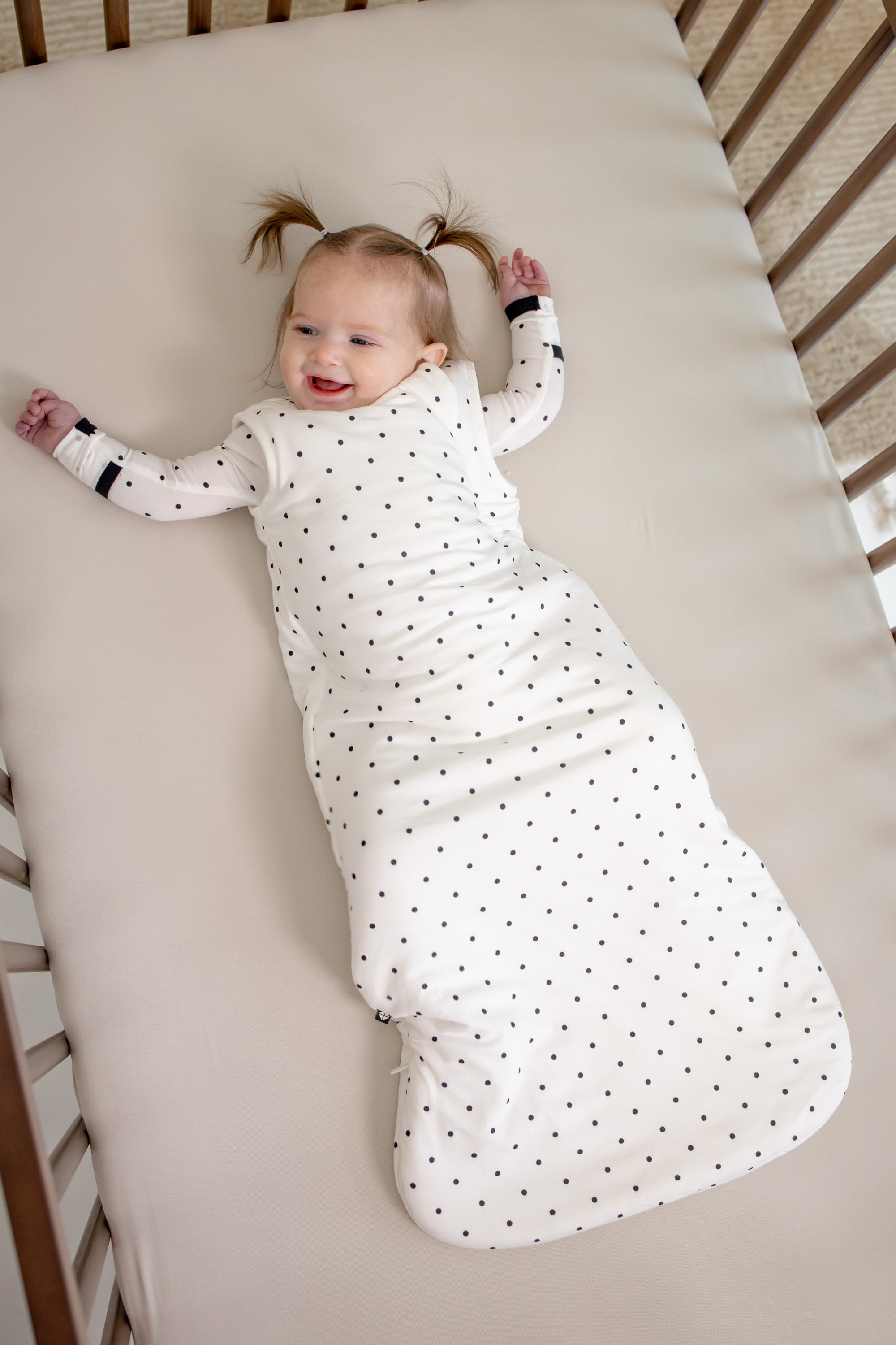 Baby lying in a crib wearing Sleep bag in Midnight Polka Dot print on a beige crib sheet