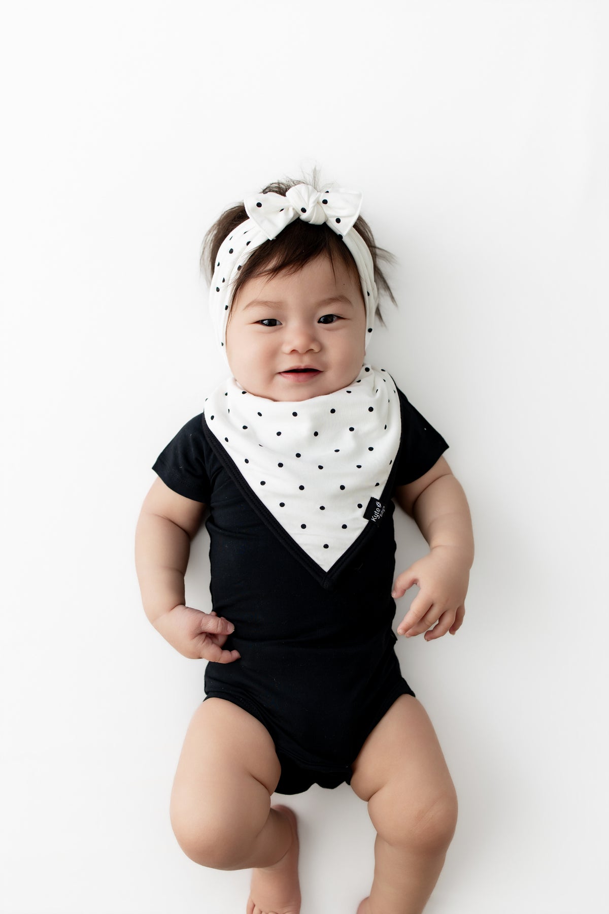 Baby wearing Midnight Polka Dot print Bib and Bow headband over a black bodysuit on a white background