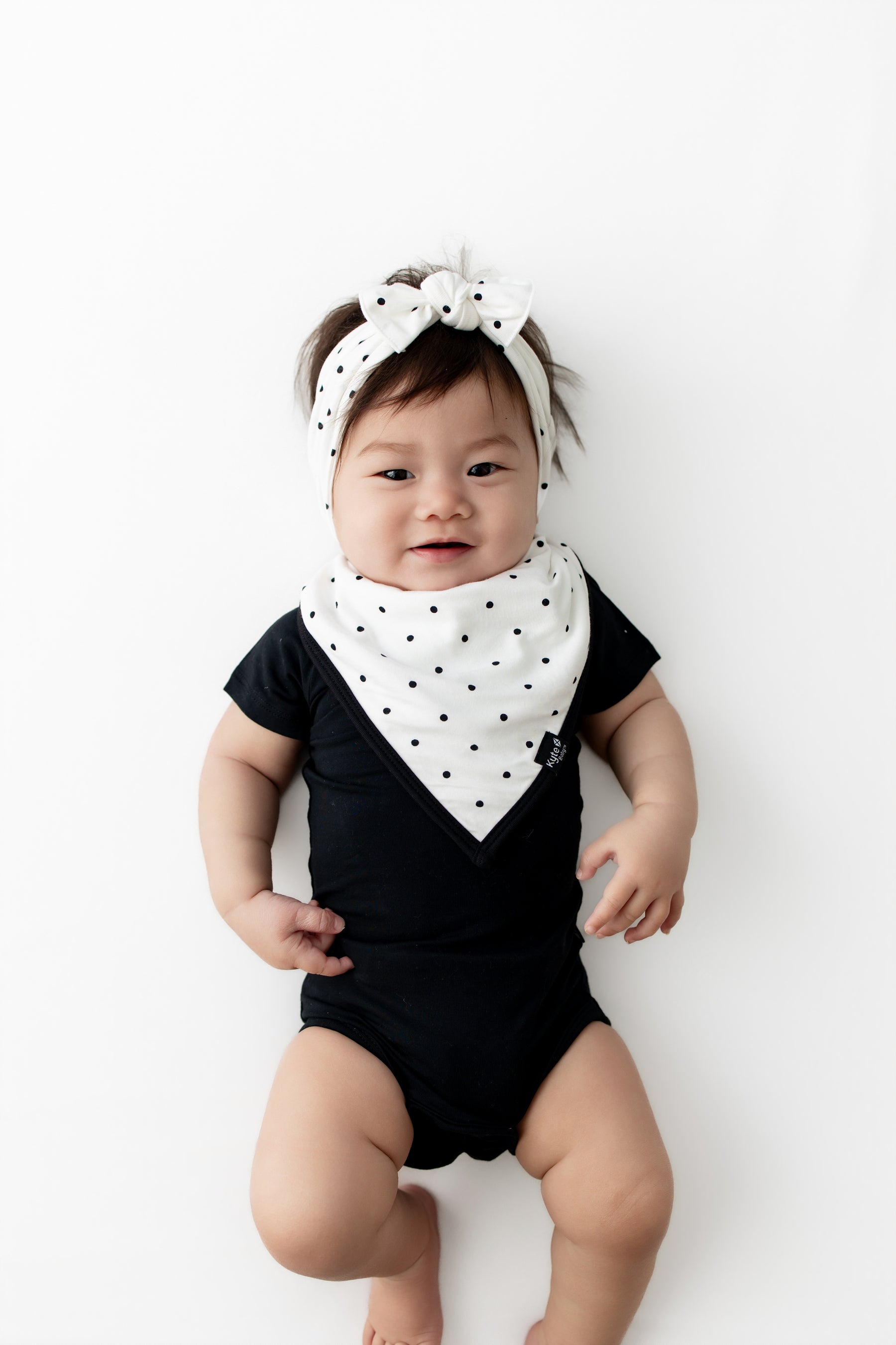 Baby wearing Midnight Polka Dot print Bib and Bow headband over a black bodysuit on a white background
