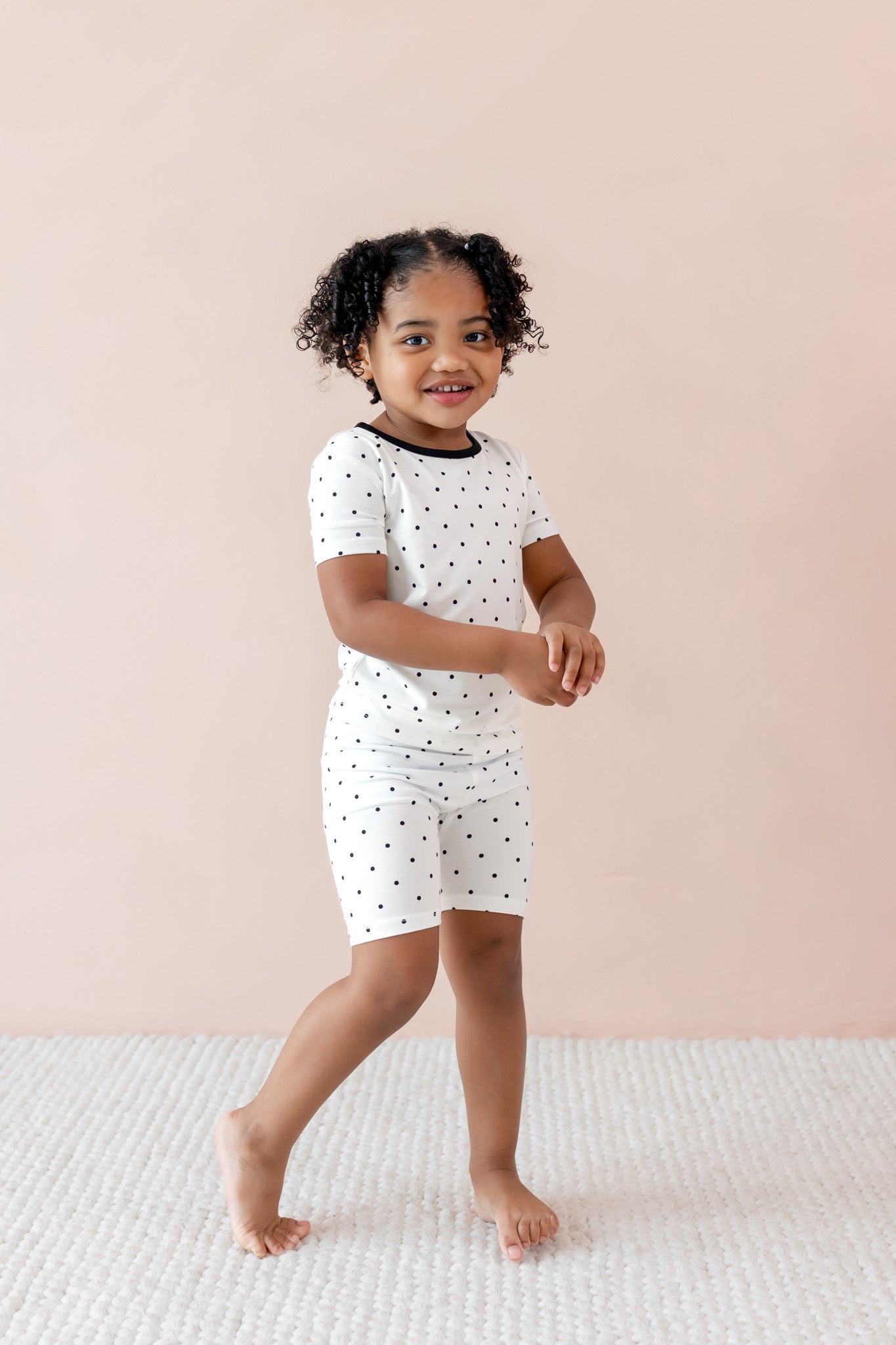 Child wearing Toddler Short Sleeve pajama set in Midnight Polka Dot print with black trim on the neck on a beige background