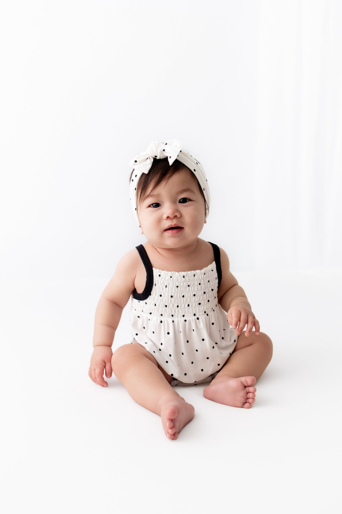 Baby wearing Midnight Polka Dot print Smocked Bubble Romper and bow headband on a white background