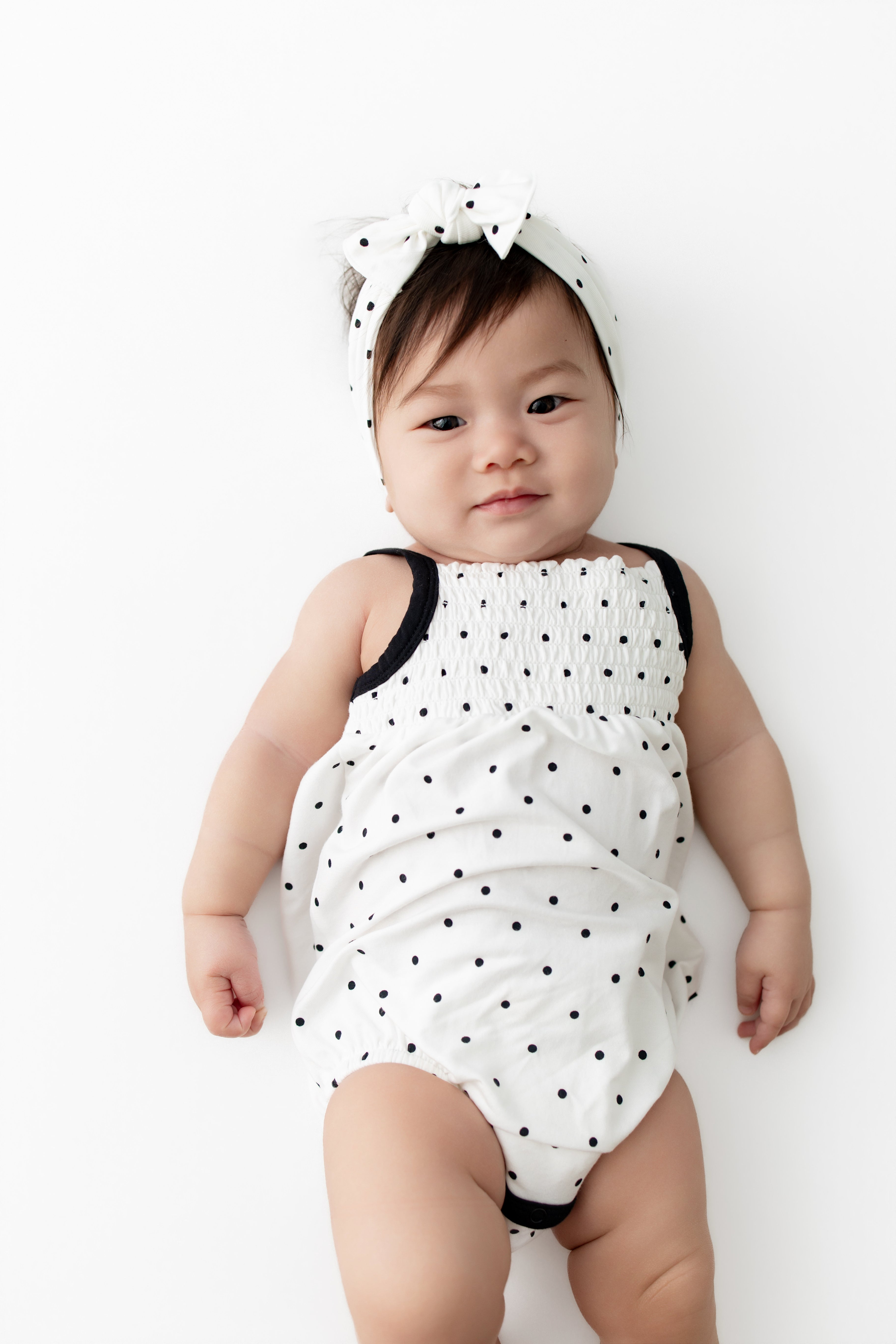Baby in Smocked Bubble Romper and bow headband in Midnight Polka Dot print on a white background