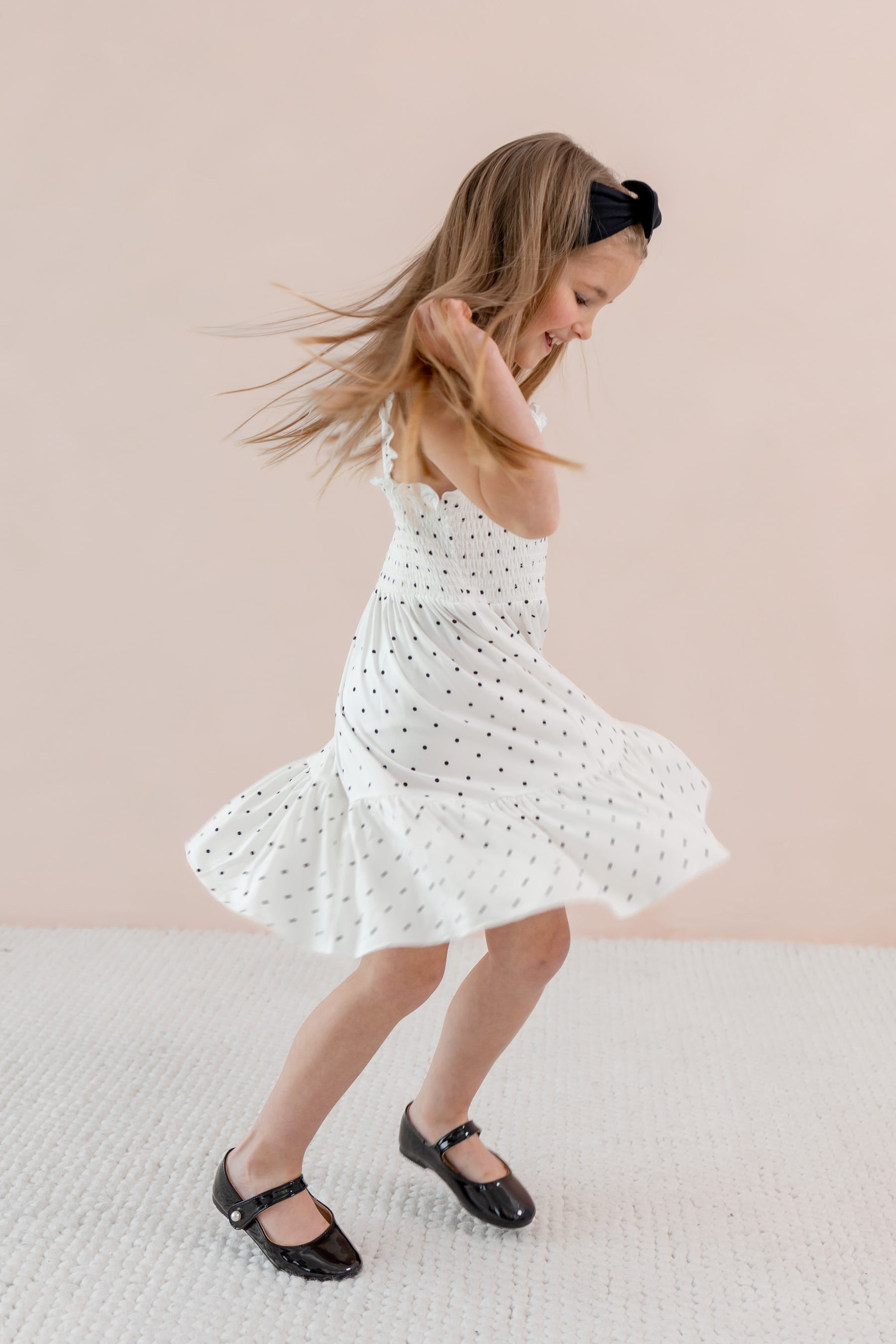 Child in Smocked Dress with Midnight Polka Dot print wearing Black bow headband and shoes on a beige background
