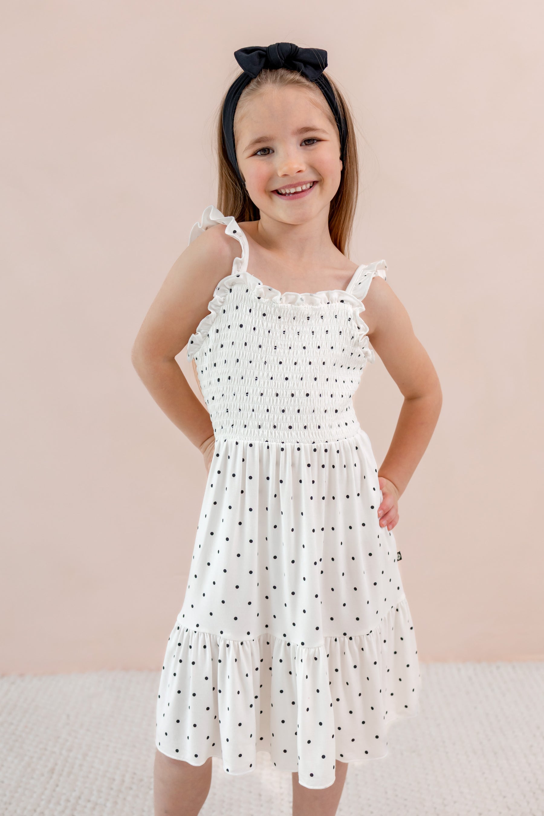 Child posing in Smocked Dress in Midnight Polka Dot print wearing Black bow headband on a beige background