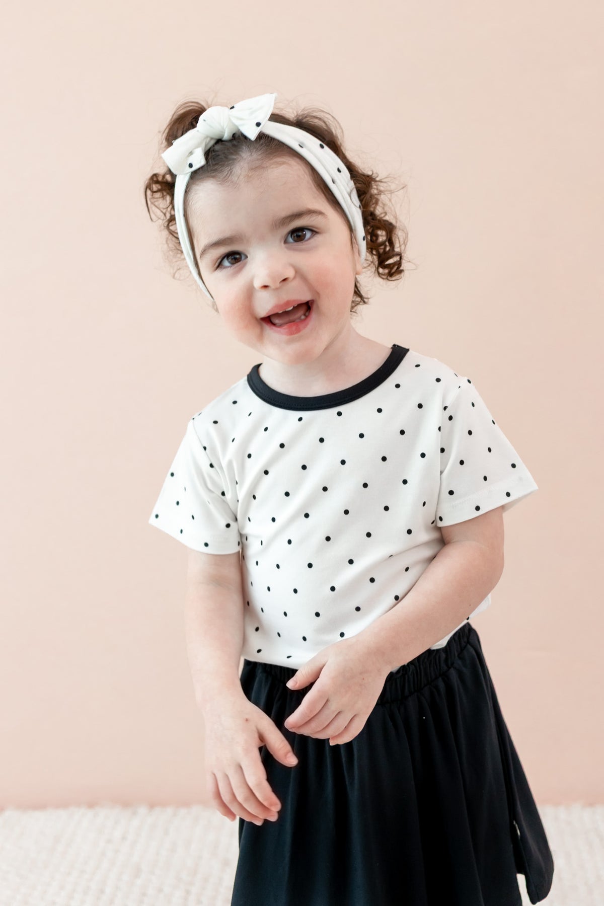 Child wearing Midnight Polka Dot print Toddler Basic Tee and bow headband over a black skort on a beige background