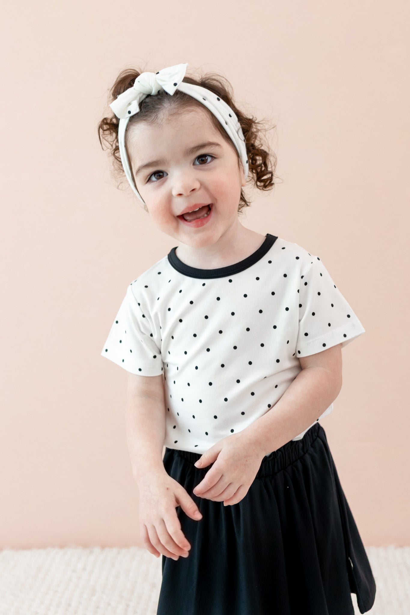 Child wearing Midnight Polka Dot print Toddler Basic Tee and bow headband over a black skort on a beige background