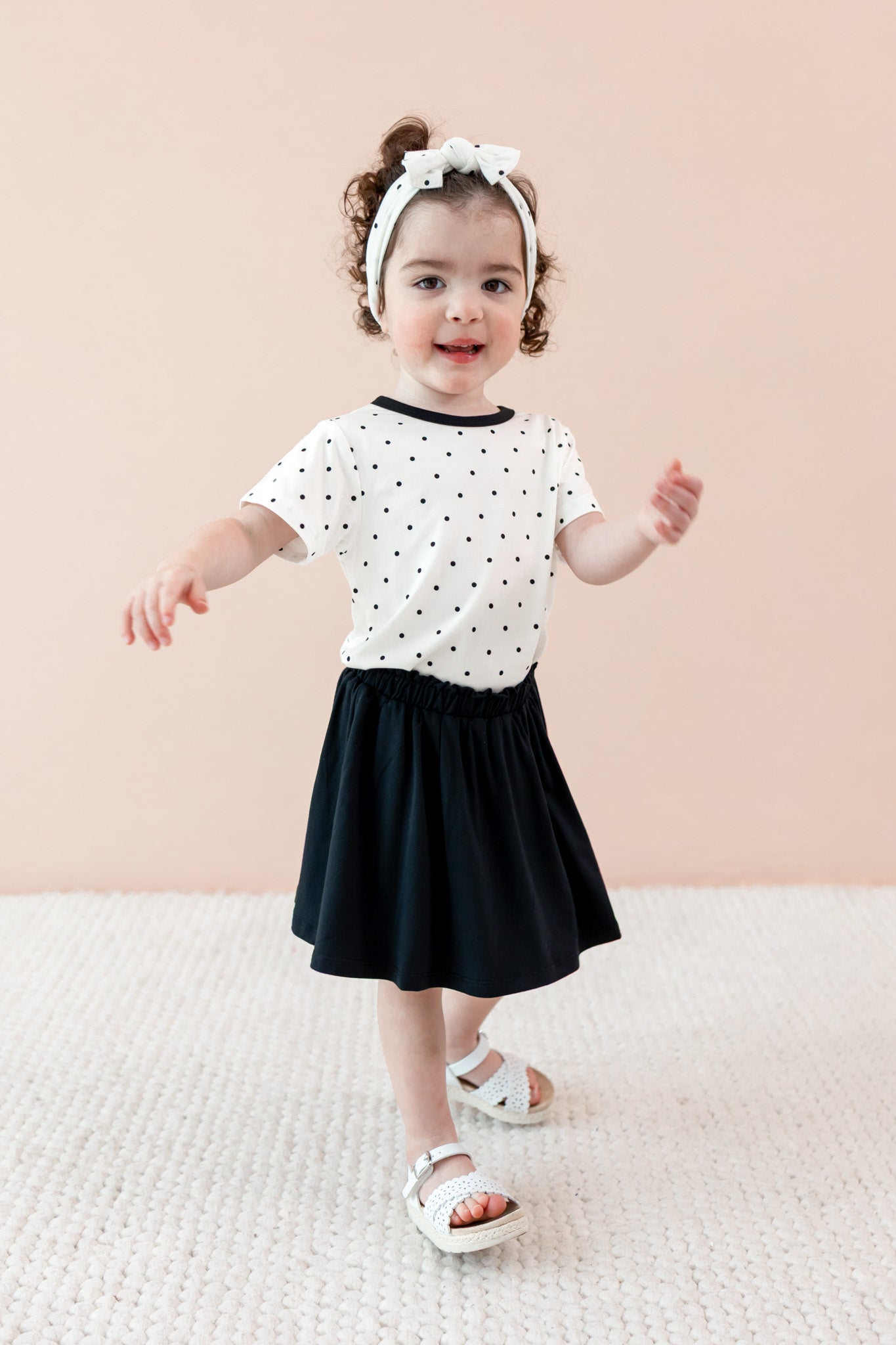 Child wearing Midnight Polka Dot print Toddler Basic Tee and bow headband over a black skort on a beige background