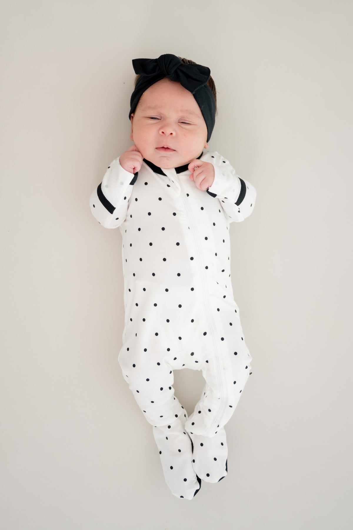 Baby sleeping in midnight polka dot print zippered footie with black bow headband on a beige background