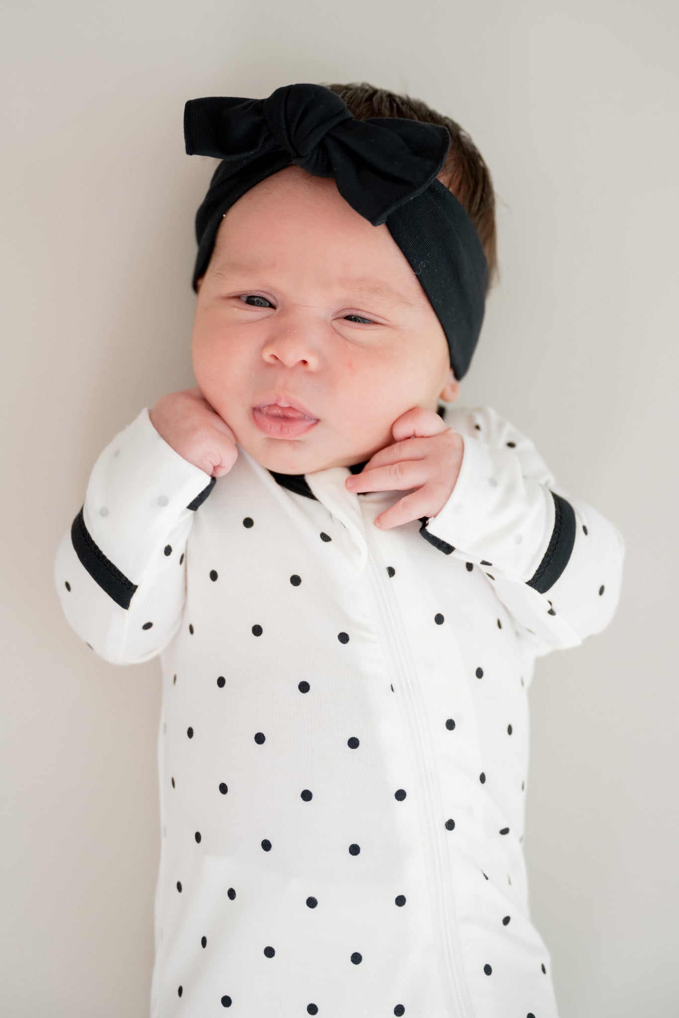 Baby wearing midnight polka dot print zippered footie with black bow headband on a beige background
