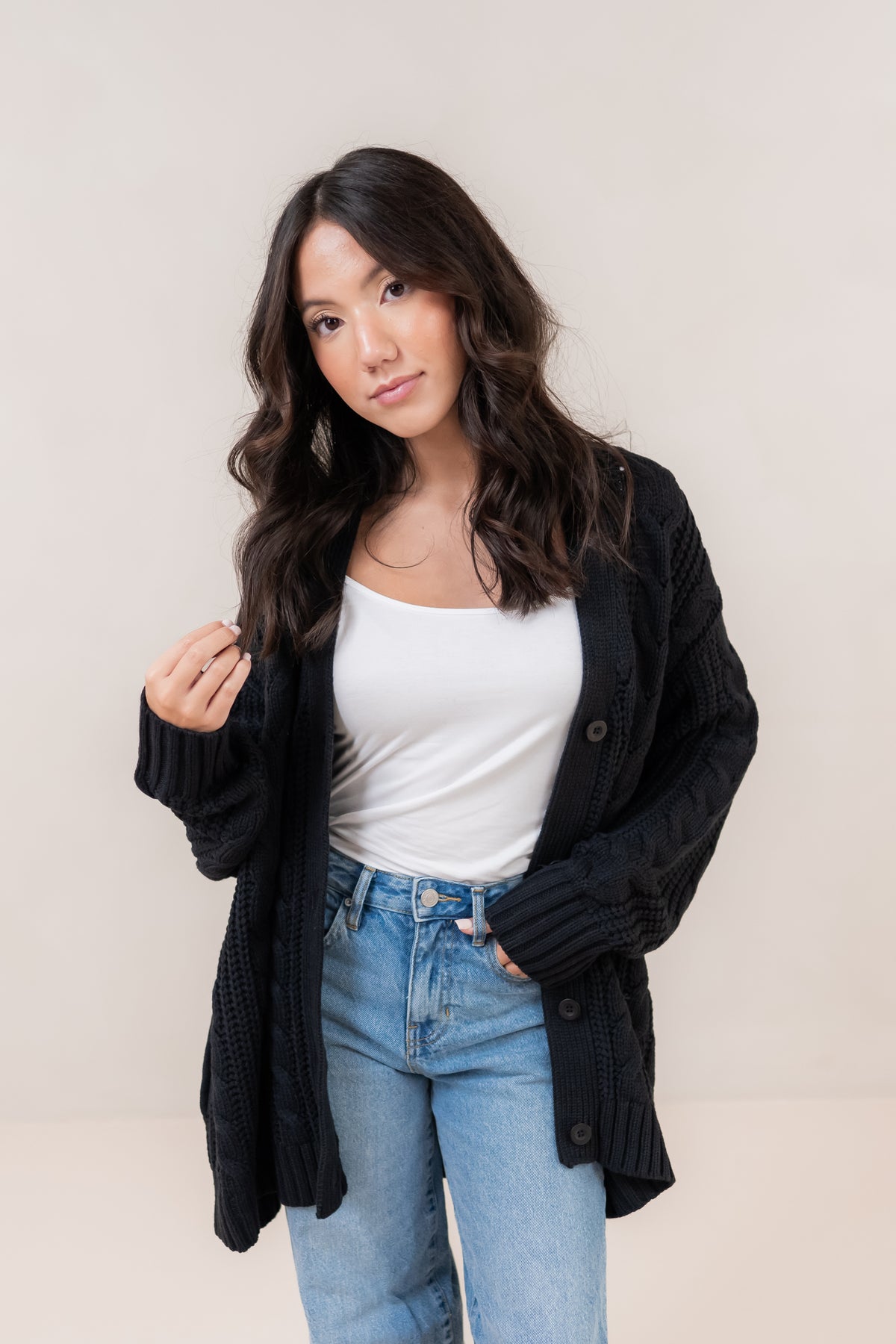 Woman wearing a black cardigan, white top, and blue jeans against a plain background