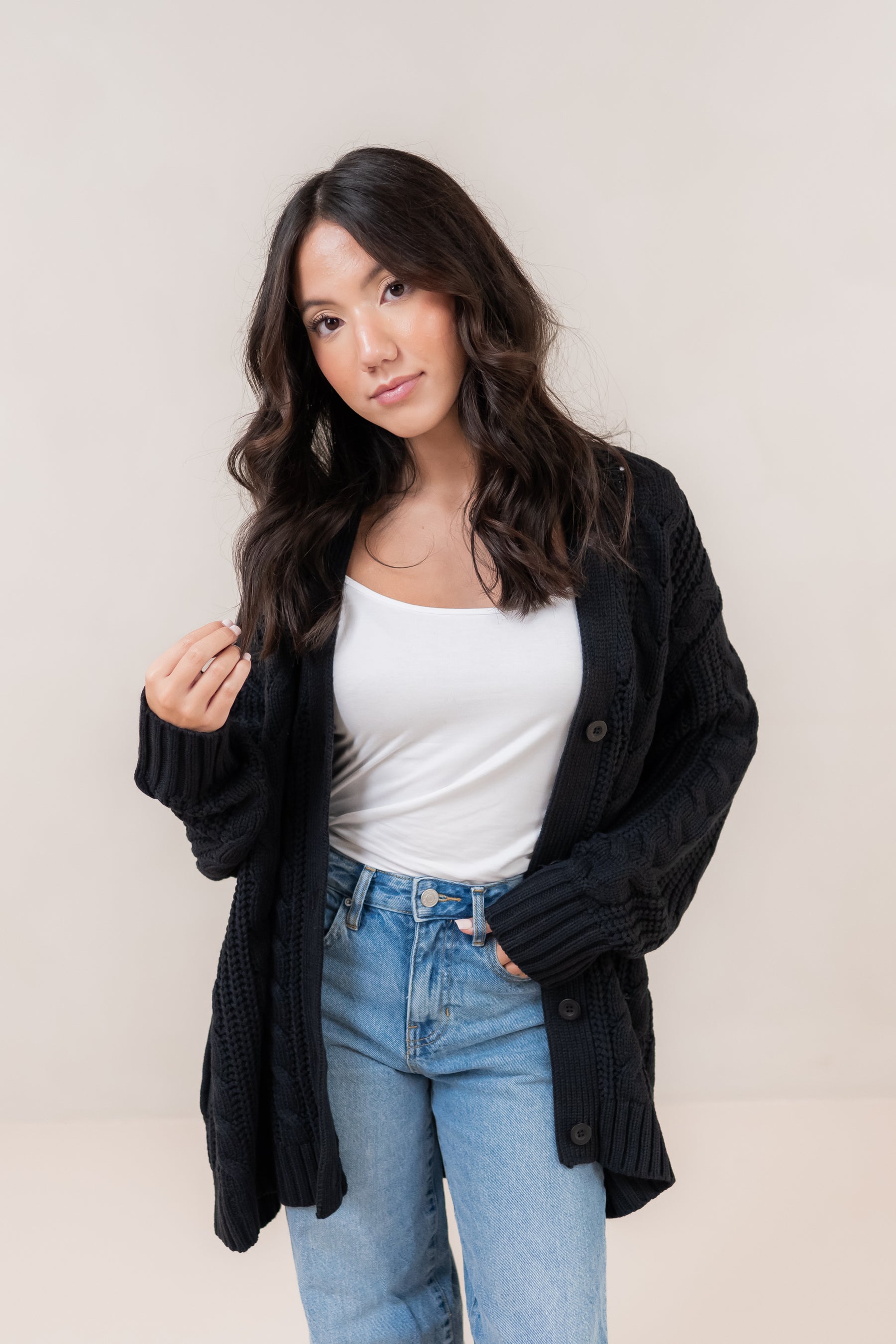Woman wearing a black cardigan, white top, and blue jeans against a plain background