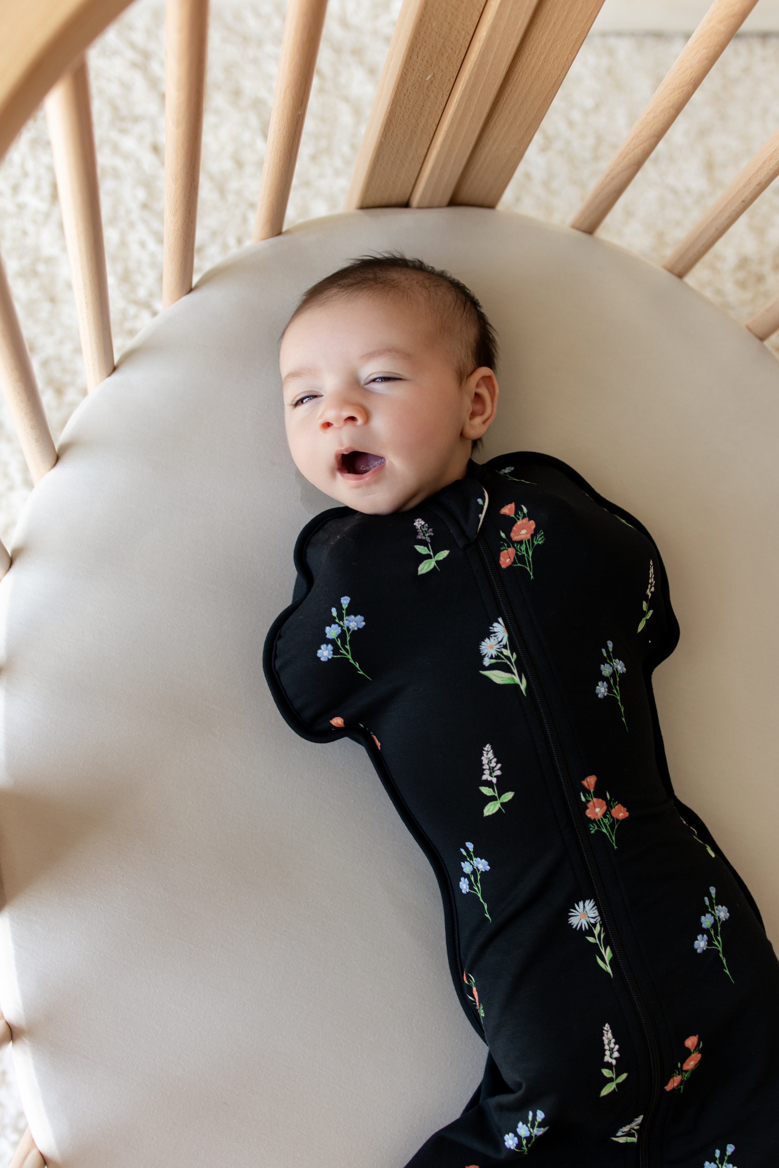 Baby yawning in a crib wearing black Swaddle Bag with Wildflower print