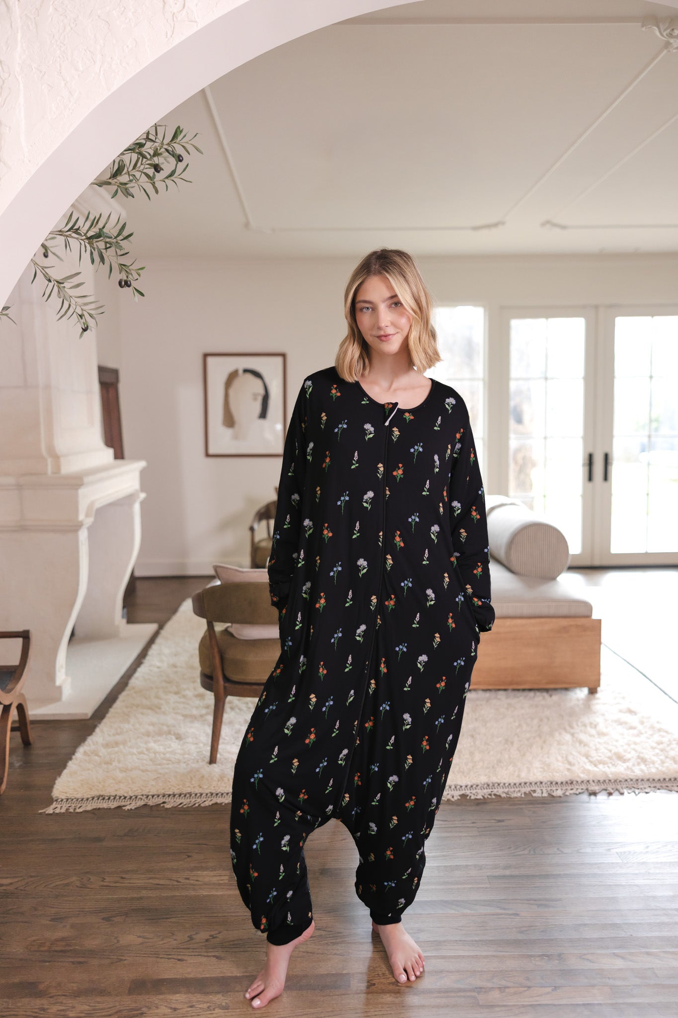 A woman stands barefoot in a bright, modern room wearing a black harem-style onesie with a small all-over floral print.