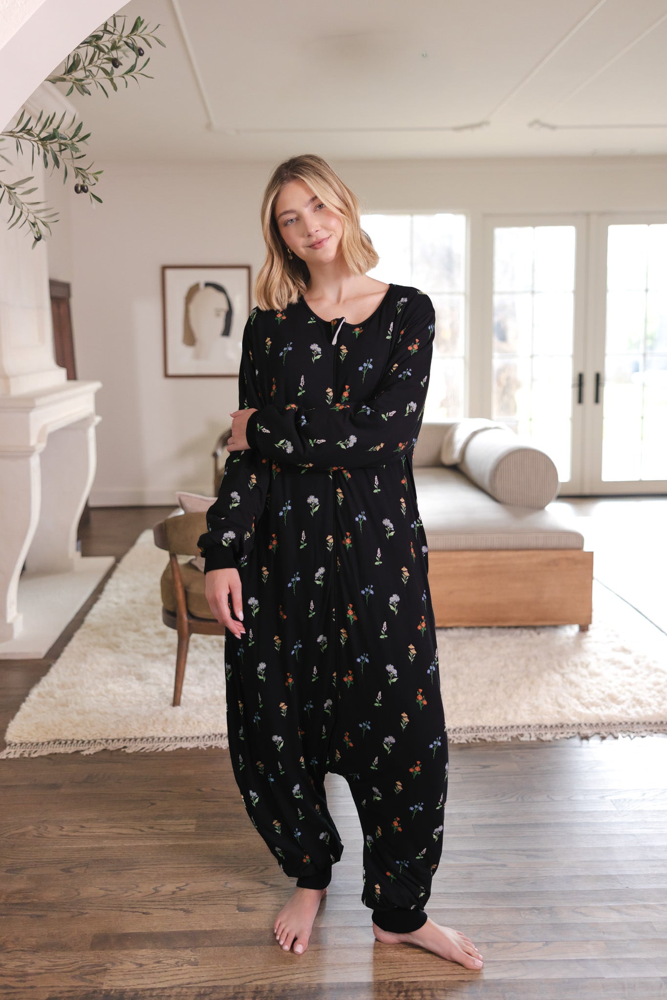 A woman modeling a black long-sleeve harem-style onesie with a small multicolor floral print, standing barefoot in a bright, modern living room.