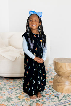 Girl posing in Sleep Bag Walker in Midnight Wildflower 1.0 print with a blue bow on head standing in a living room