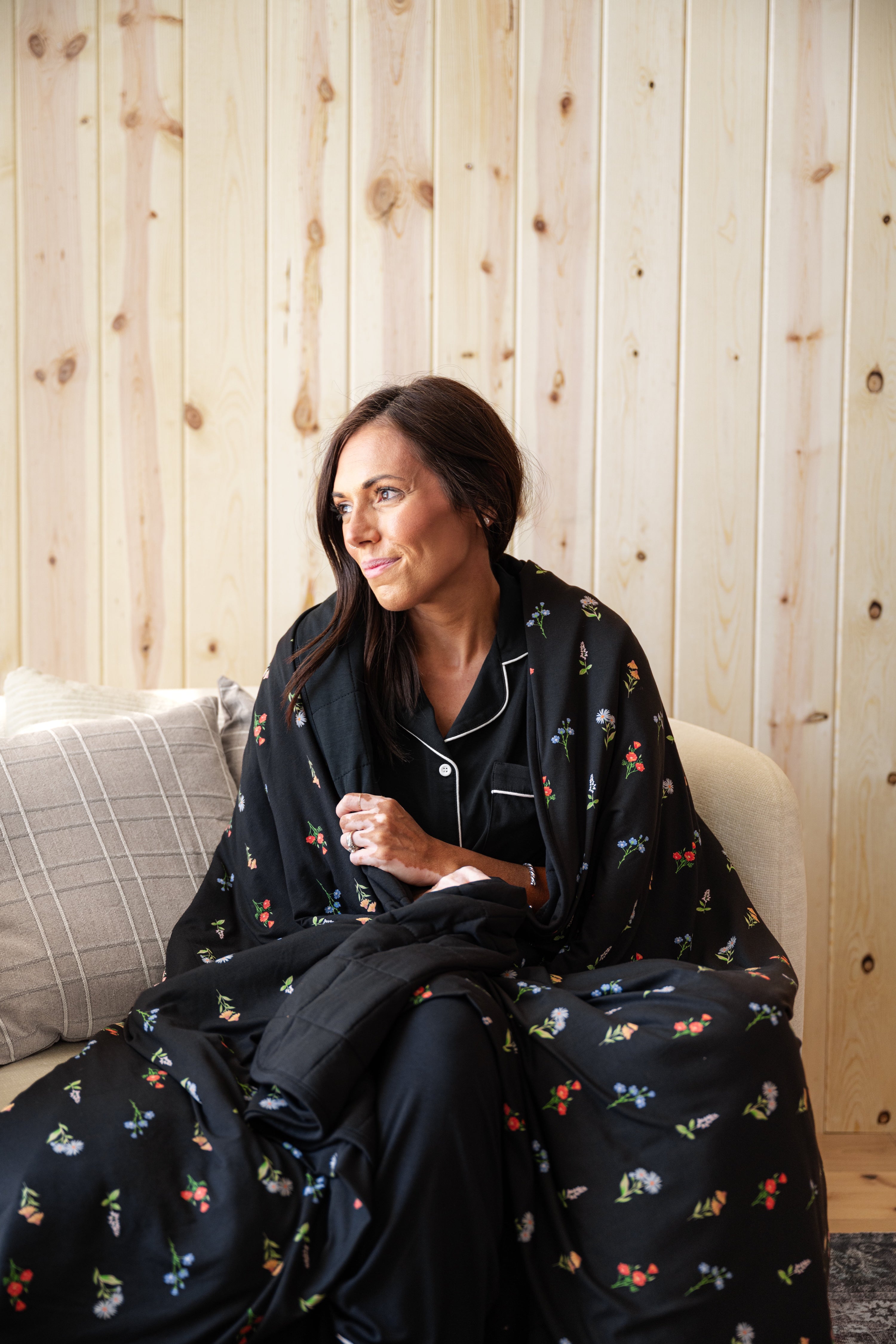 A woman sits on a light-colored sofa wearing a black pajama set with a small multicolor floral print and white piping. She is wrapped in a matching black blanket with the floral pattern.