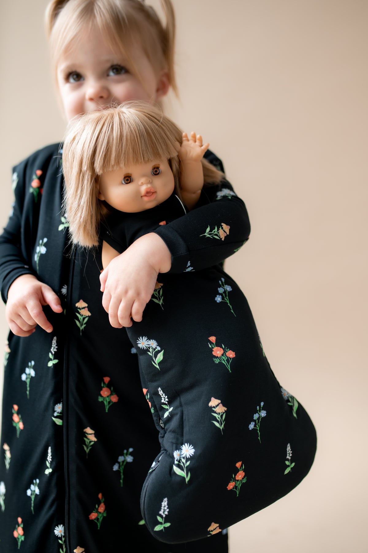 Girl holding a baby doll sleep bag in midnight wildflower while also wearing Sleep Bag Walker in Midnight Wildflower 2.5 print on a beige background
