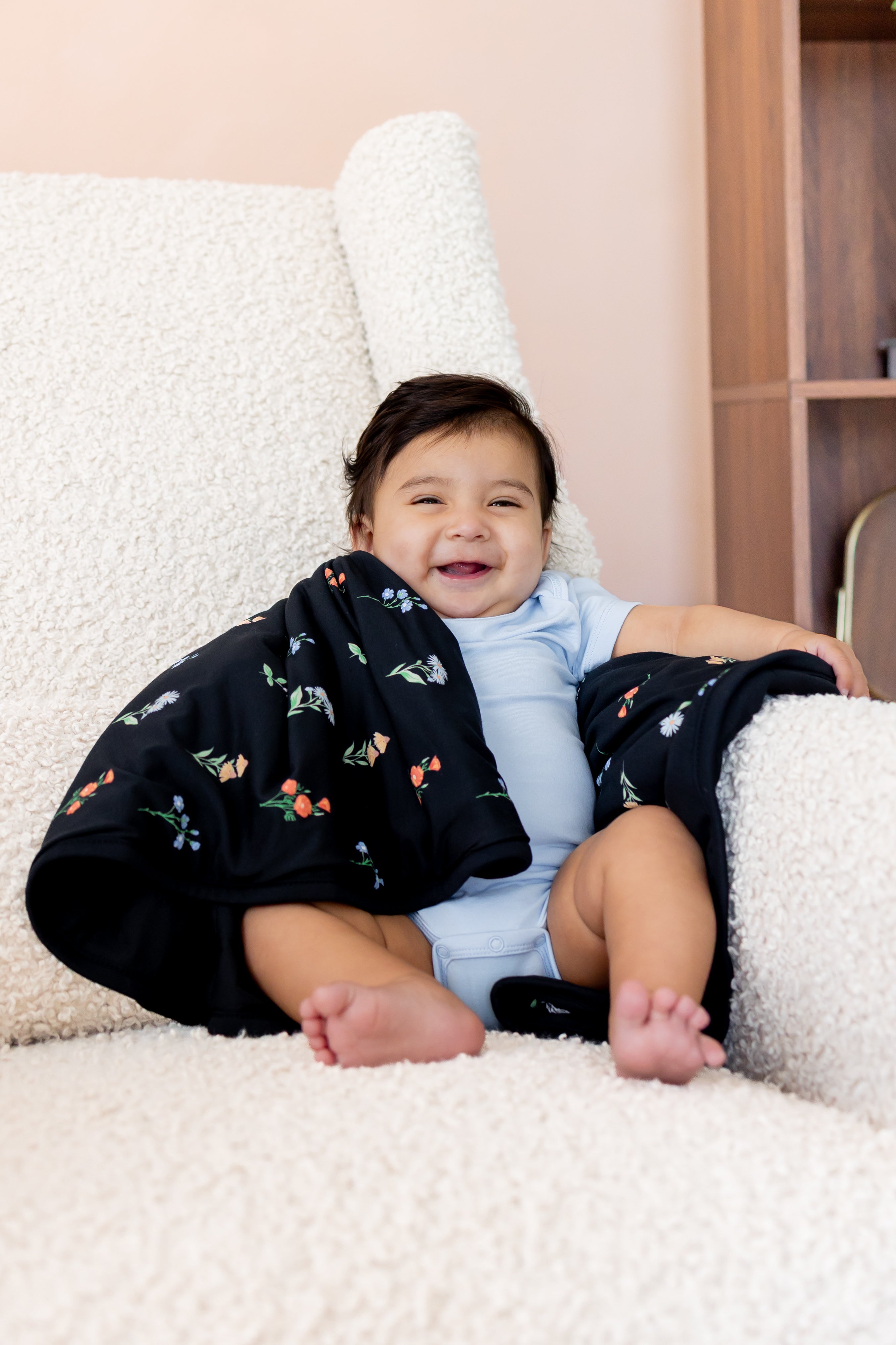 Baby smiling on white sofa wearing a blue bodysuit with Baby Blanket in Midnight Wildflower 1.0 print