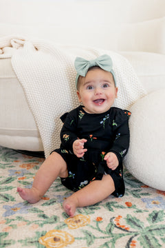 Baby sitting against a white couch smiling in Long Sleeve Twirl Bodysuit Dress in Midnight Wildflower print and a blue head bow