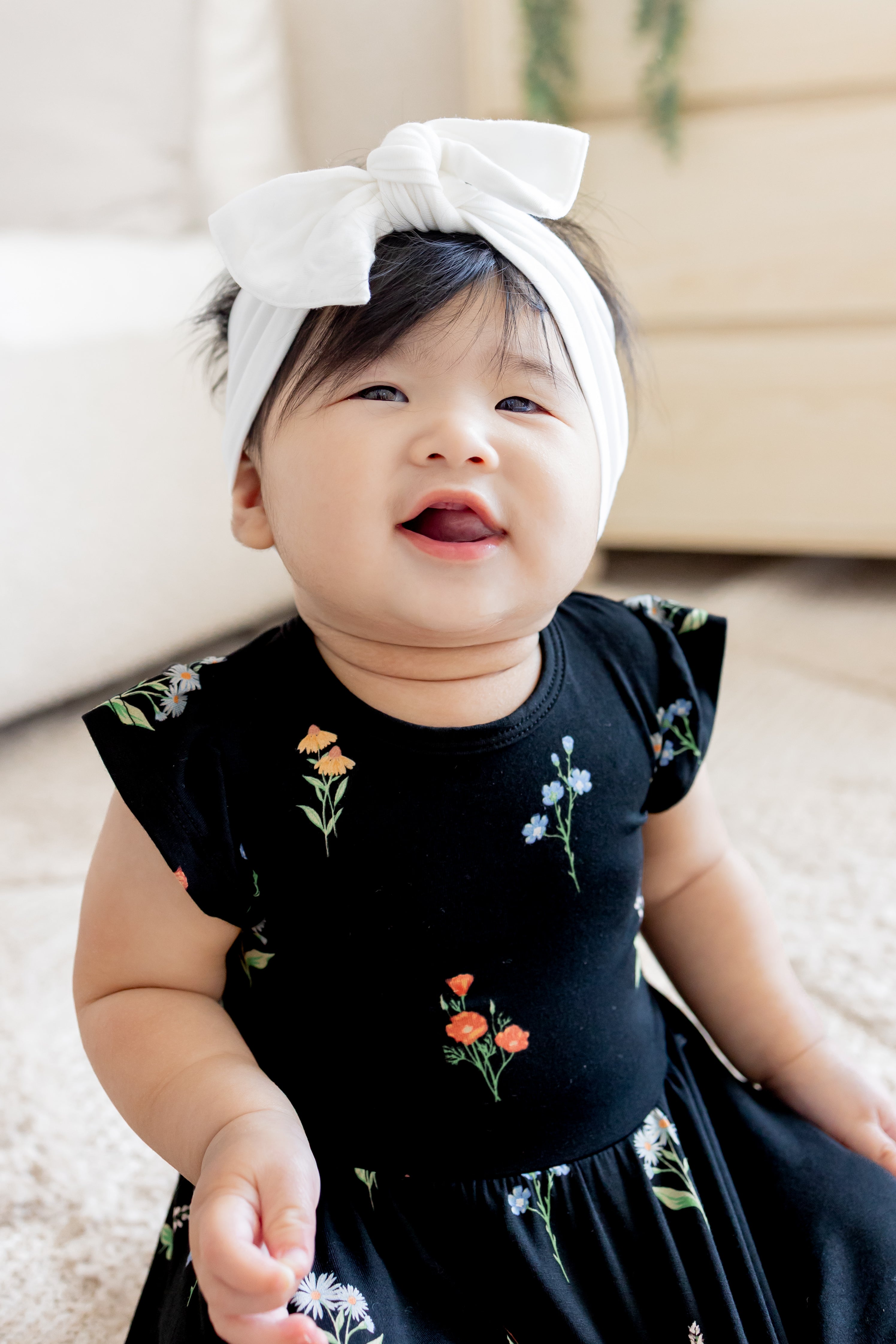 Baby smiling in Twirl Bodysuit Dress in Midnight Wildflower print wearing a white head bow on a white carpet