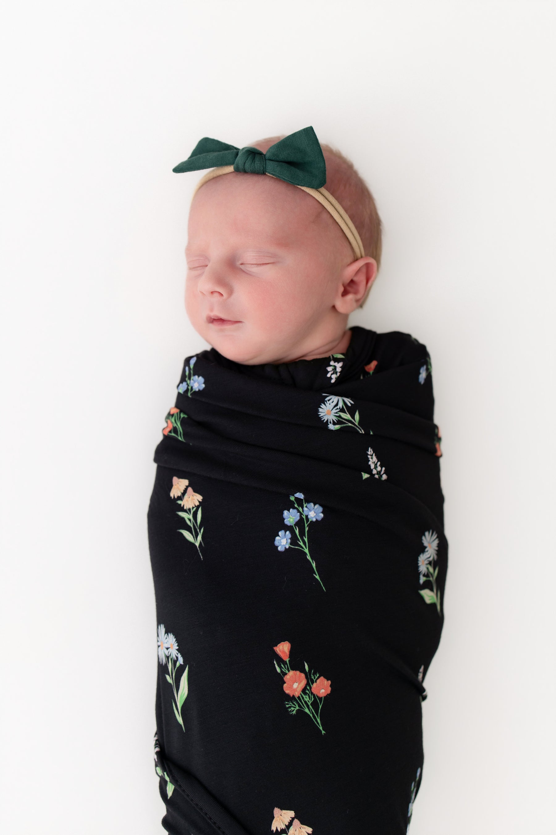 Baby in Swaddle Blanket in Midnight Wildflower print and green head bow on a white background