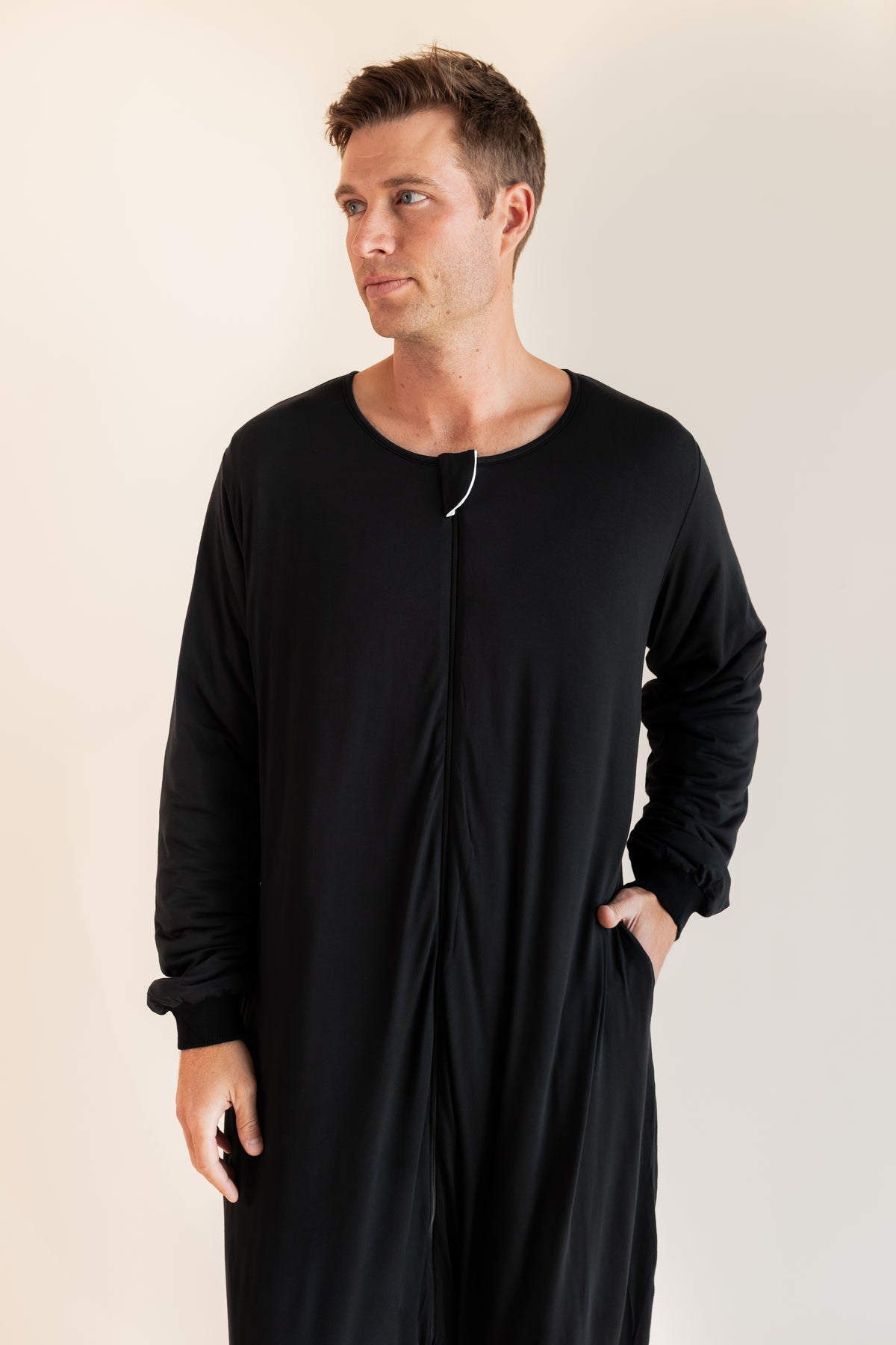 A man wearing a black long-sleeve sleep sack with a front zipper and side pockets, standing against a plain light beige studio background.