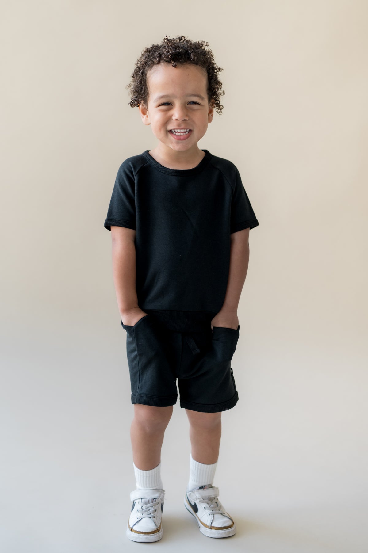 Young boy standing in front of a light beige surface wearing the Short Sleeve Jogger Set in Midnight with his hands in the pocket of the shorts