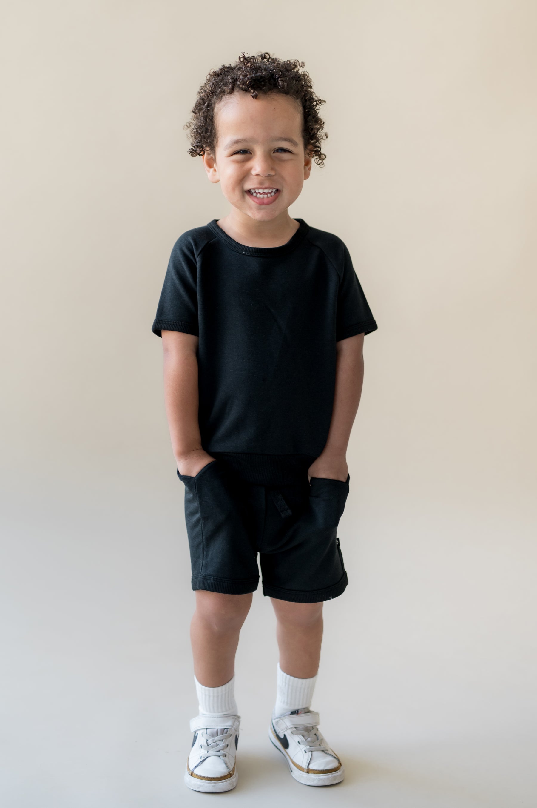 Young boy standing in front of a light beige surface wearing the Short Sleeve Jogger Set in Midnight with his hands in the pocket of the shorts