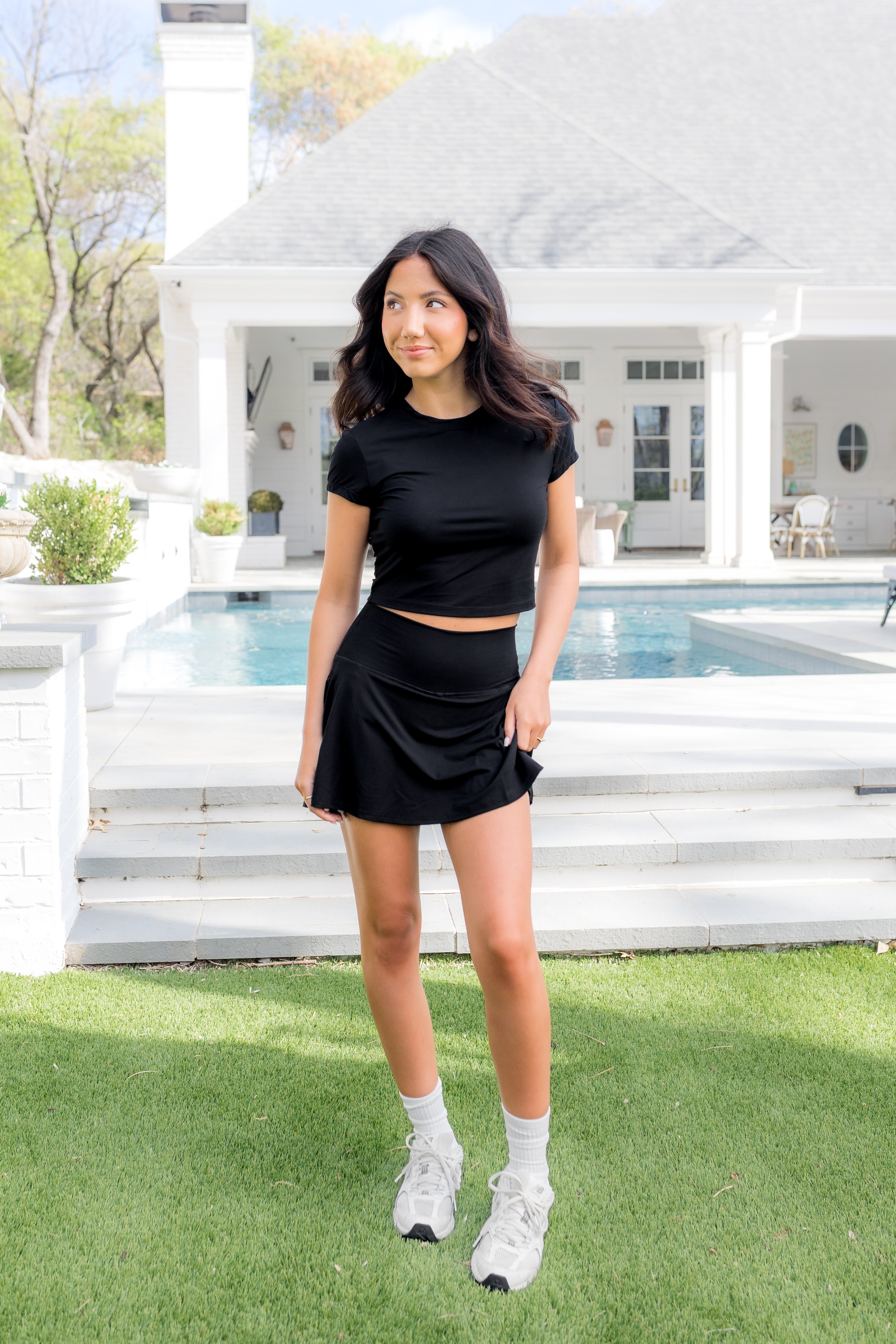 Female model posing in the grass in front of a pool wearing the Bamboo Flex Baby Tee in Midnight with matching skort