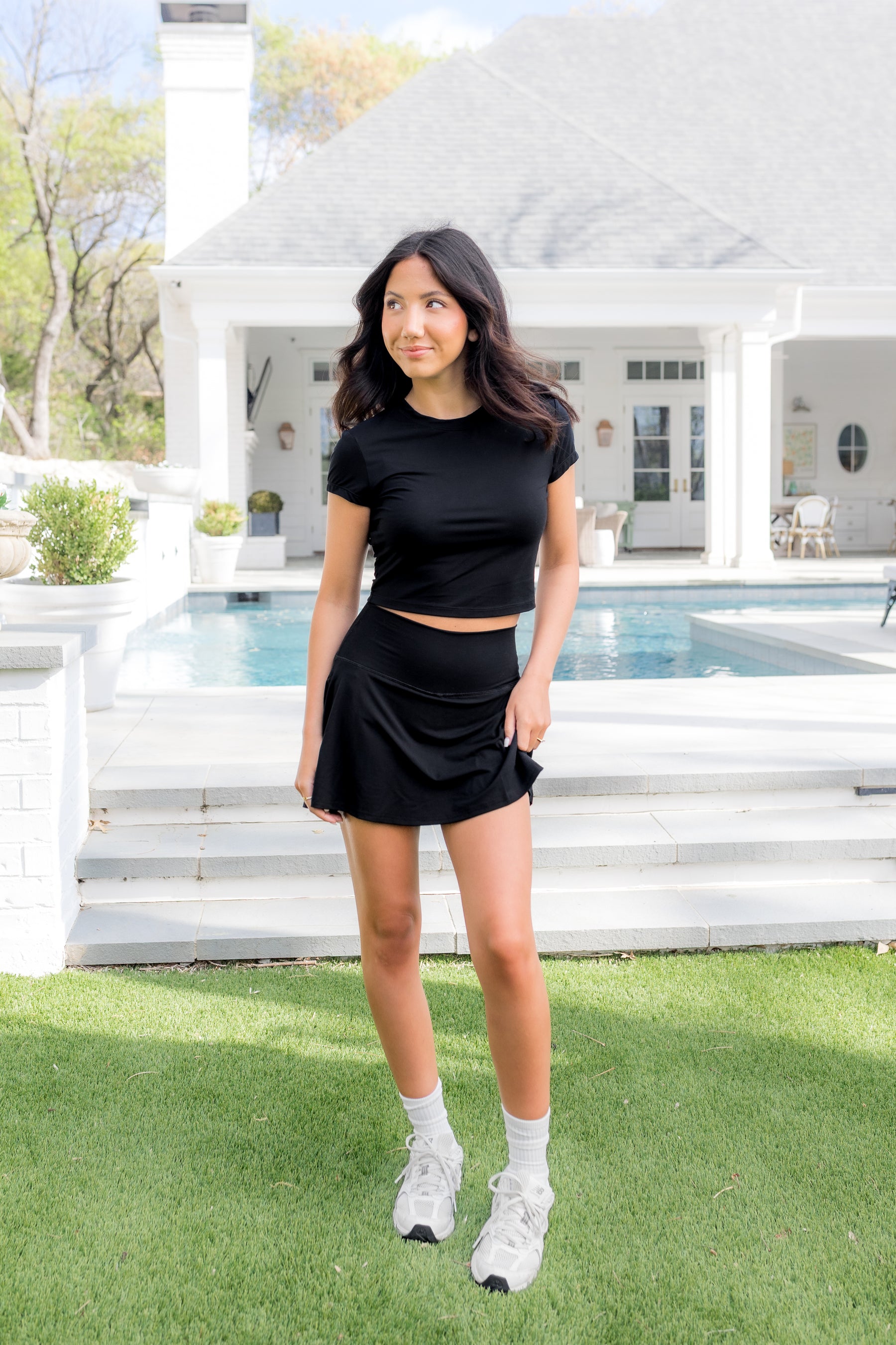 Female model posing in the grass in front of a pool wearing the Bamboo Flex Baby Tee in Midnight with matching skort