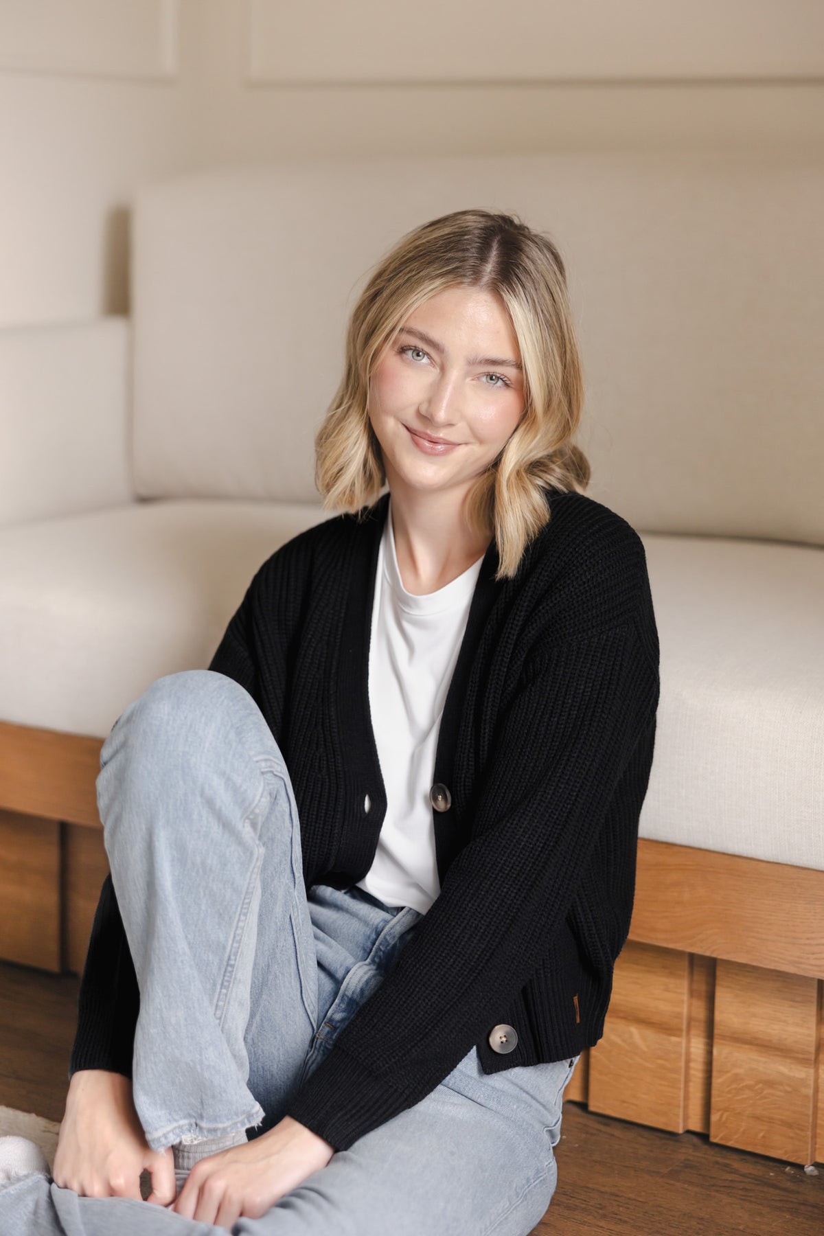 Modal smiling in Chunky Knit Women's Cropped Cardigan in Midnight over white T-shirt and Blue jeans sitting against a white sofa