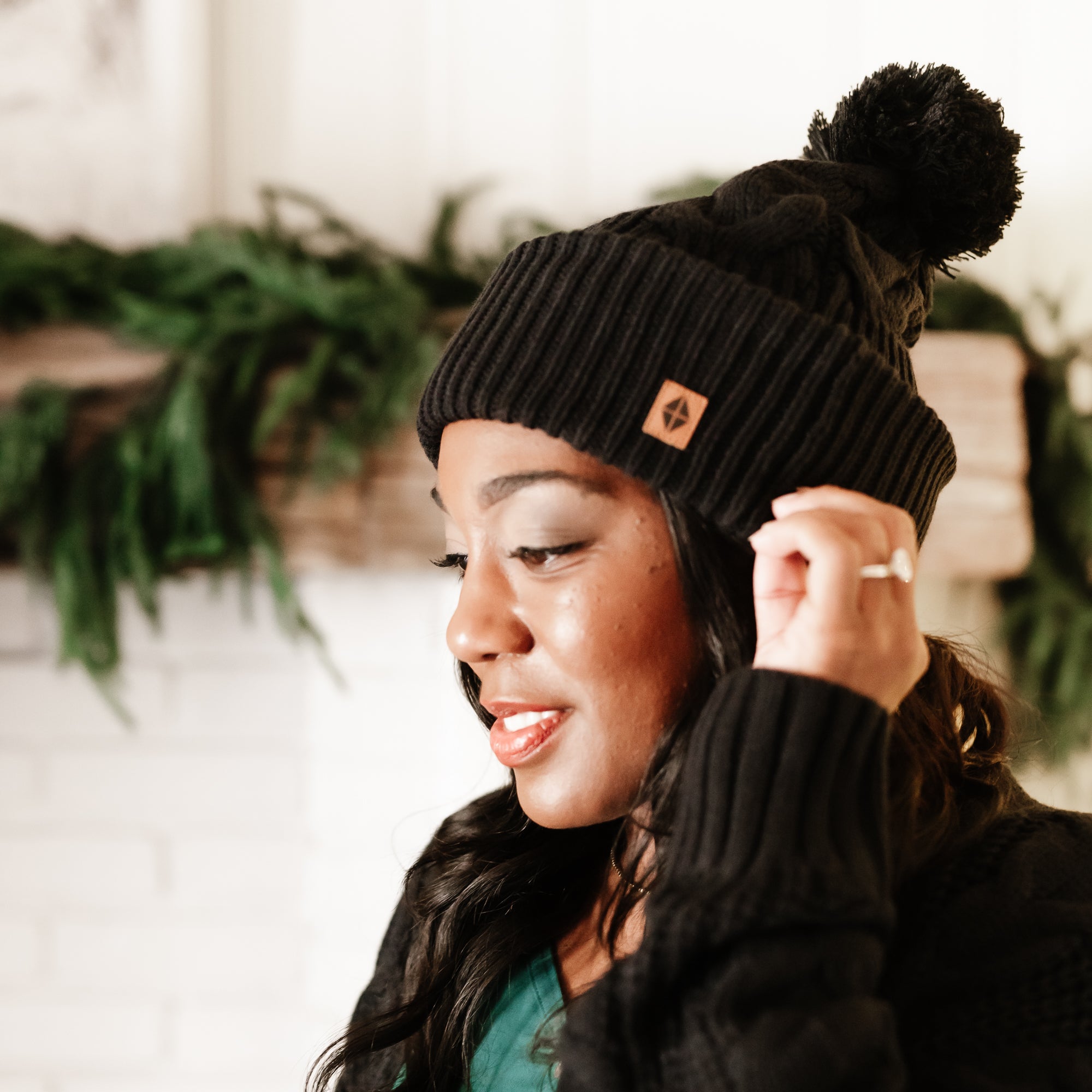 Close up of the Cable Knit Adult Pom Beanie in Midnight shown on a model with a white fireplace with greenery on the mantel in the background