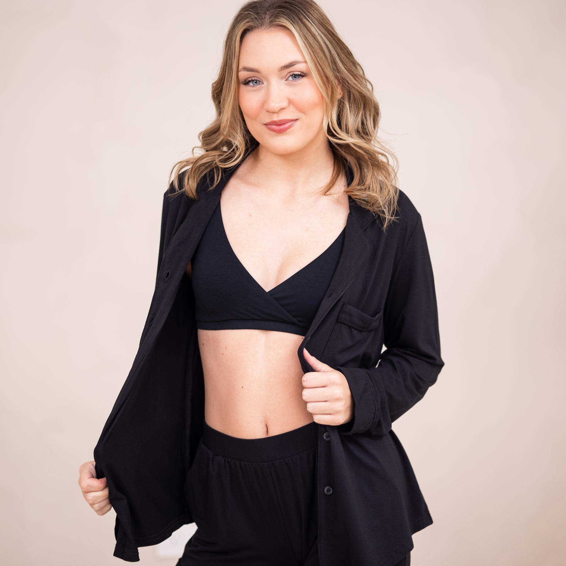 Female standing in front of a light beige wall wearing the Women's Cross Front Bralette in Midnight underneath the women's long sleeve pajama set in midnight unbuttoned