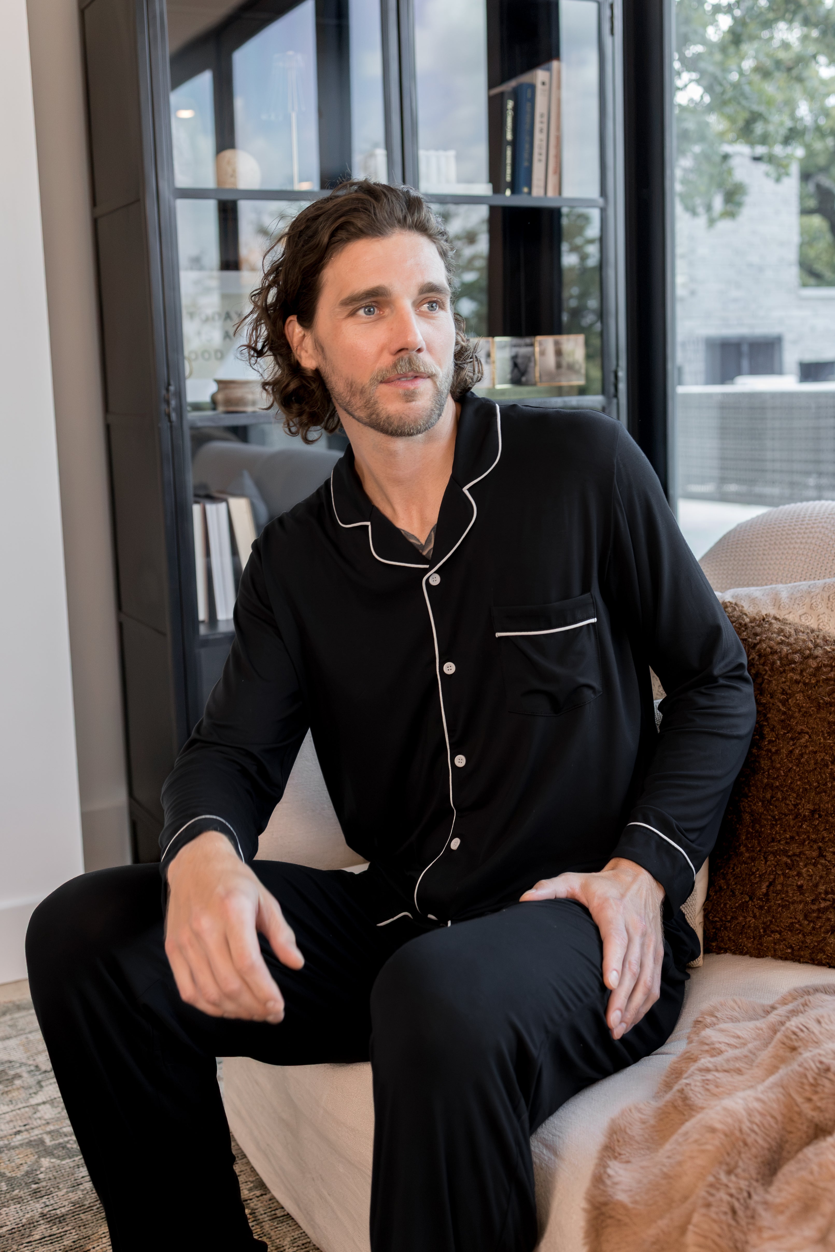 A man wearing a black long-sleeve pajama set with white piping sits on a light-colored sofa in a modern living room setting.