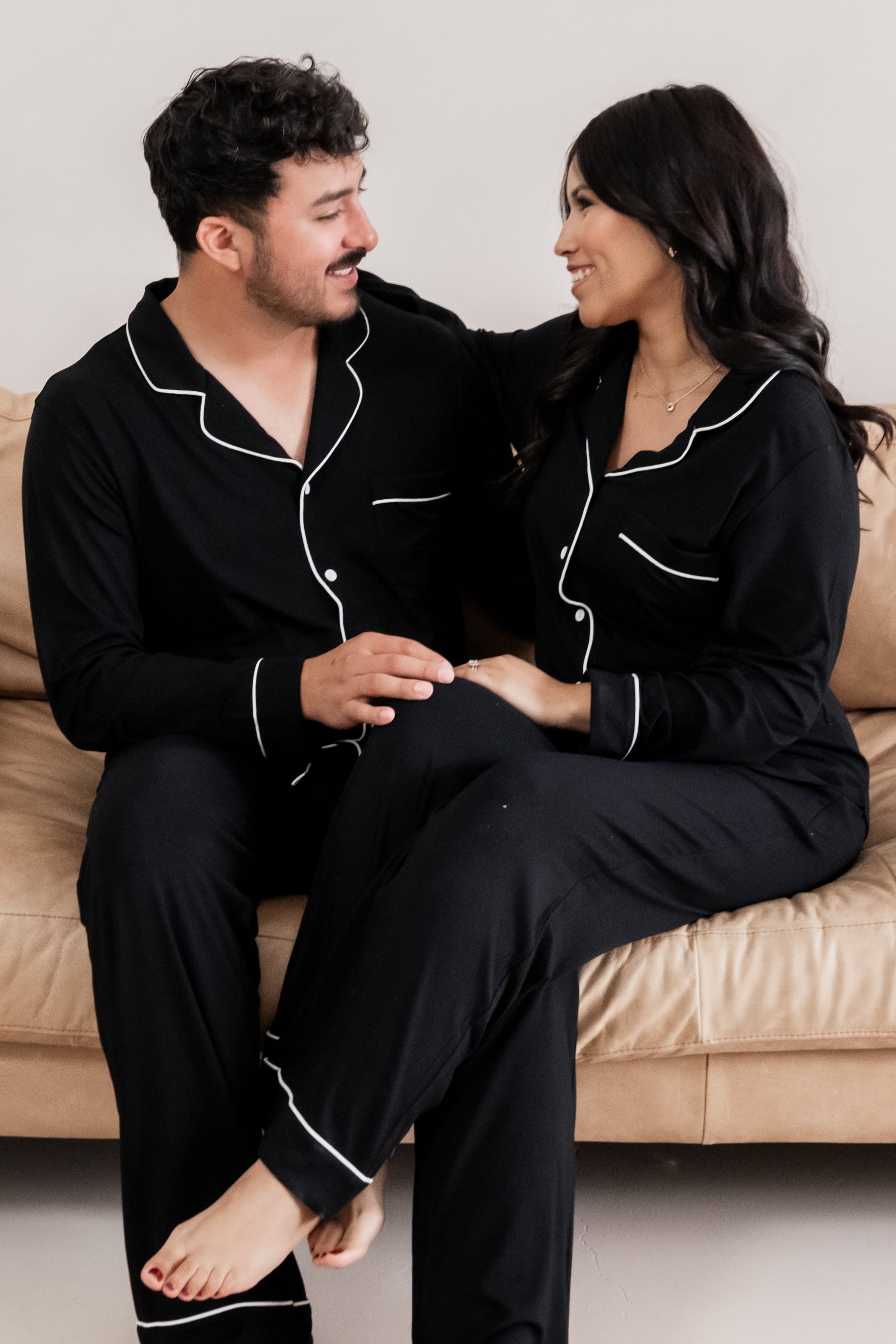 Husband and wife sitting on a couch wearing matching long sleeve midnight pajamas with white trim