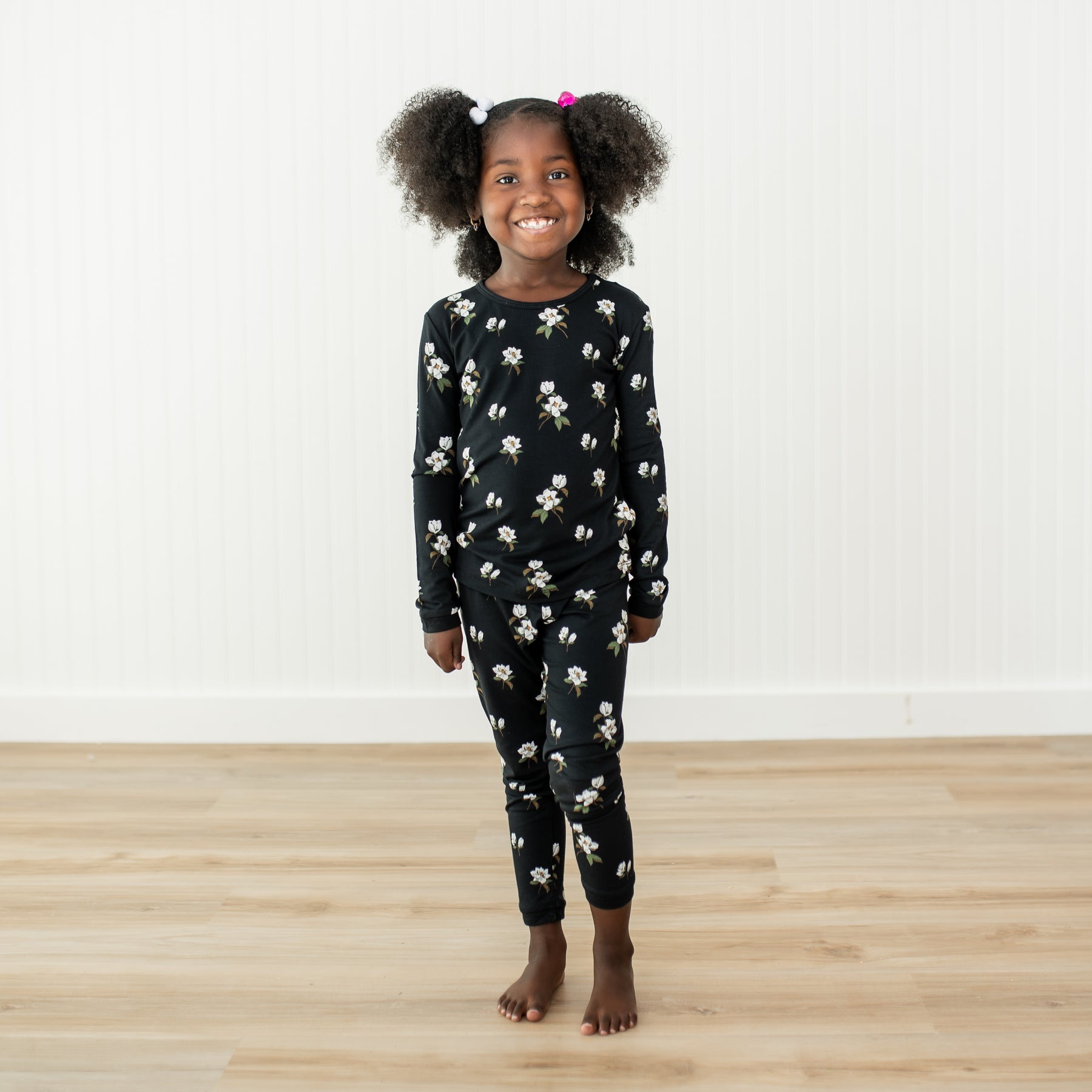 Child modeling long sleeve black pajamas with a white magnolia floral print on a wooden floor and white wall backdrop