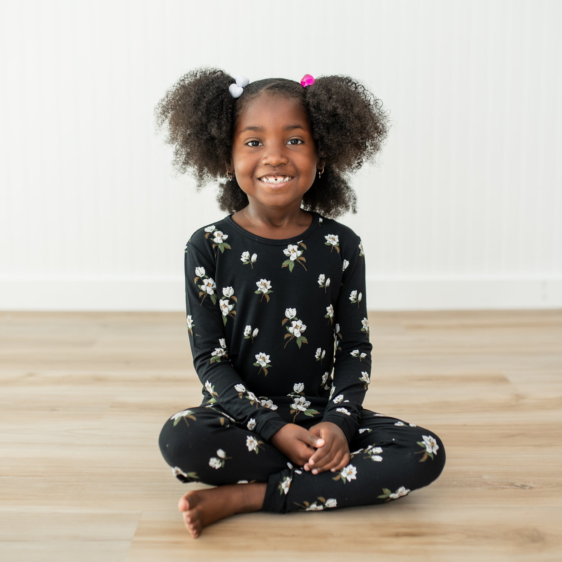 Young girl sitting on a wooden floor wearing a black floral pajama set.
