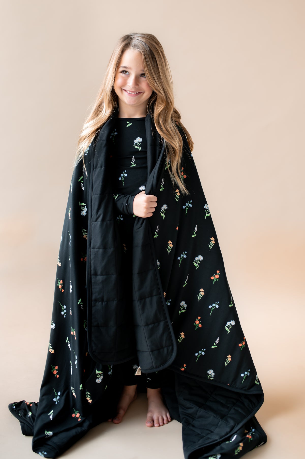 Girl standing wrapped in Youth Blanket in Midnight Wildflower 2.5 print wearing a similar print pajamas on a beige background